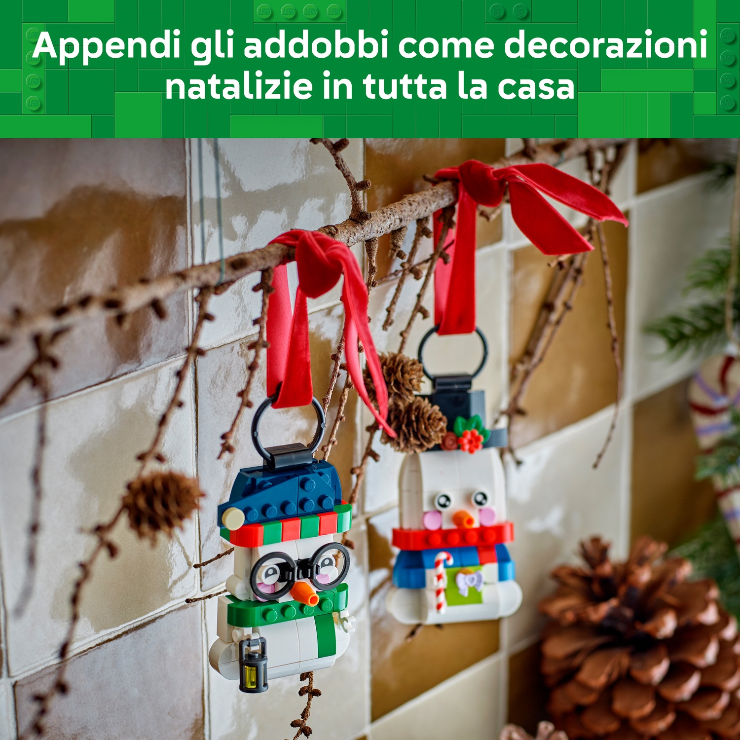 Due ornamenti di pupazzi di neve LEGO pendono da nastri rossi sui rami contro una parete piastrellata.