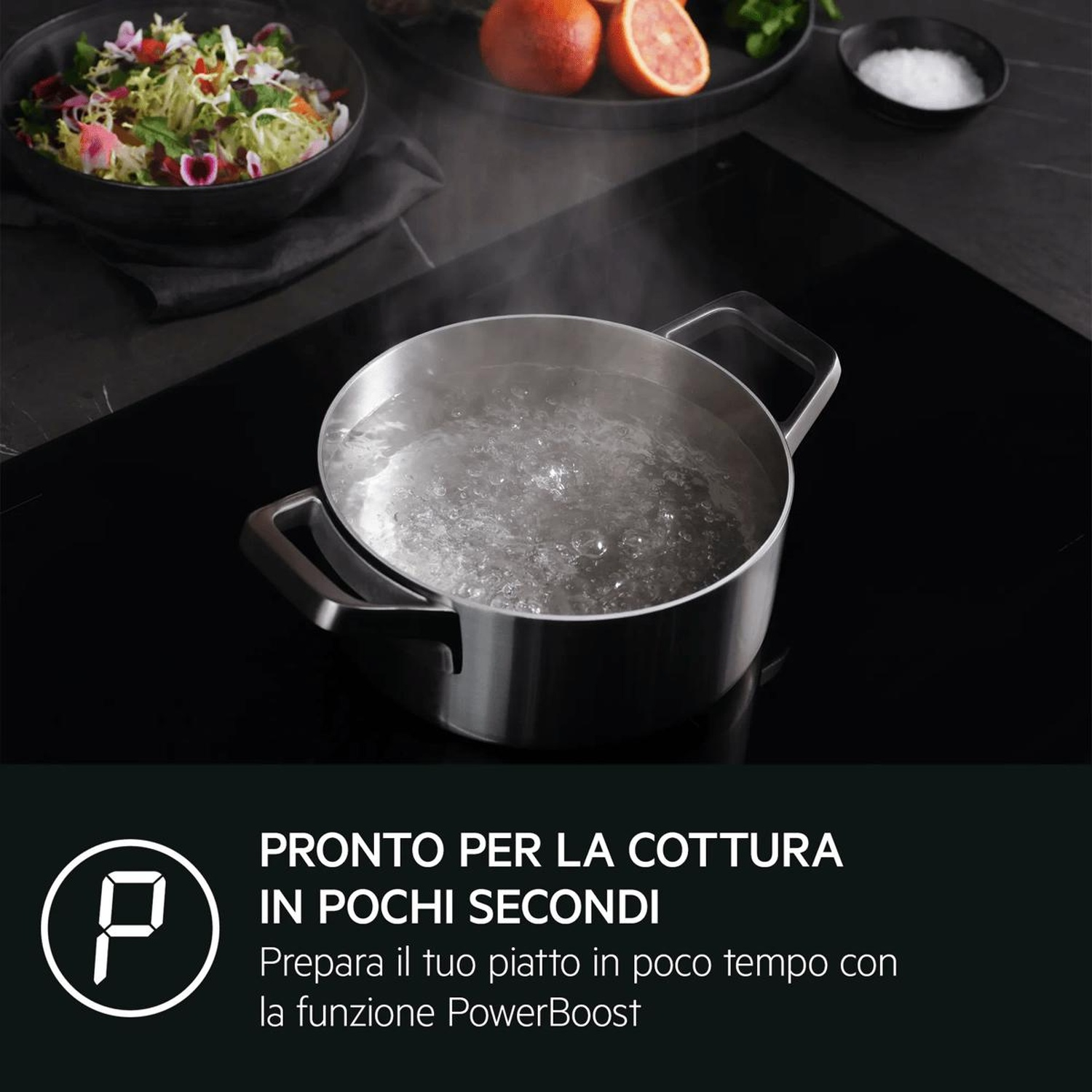 Una pentola argentata con acqua bollente su un piano cottura a induzione nero, con una ciotola di insalata sullo sfondo.