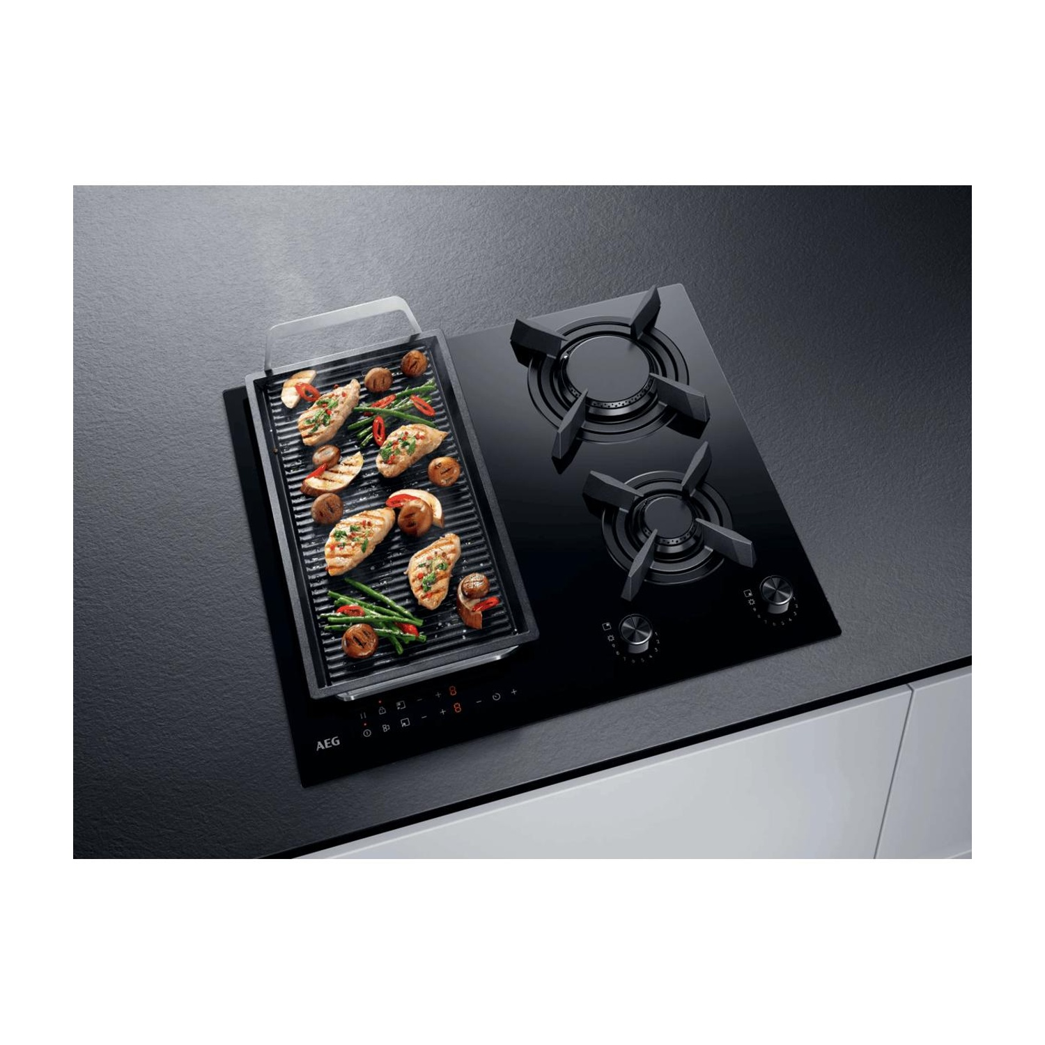 Piano cottura nero AEG con piastra grill, pollo, funghi e verdure su un piano di lavoro scuro.