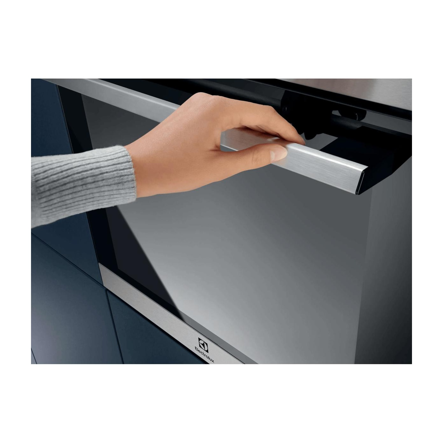 Una mano con un maglione grigio apre la maniglia in acciaio inox di un forno nero e argento.