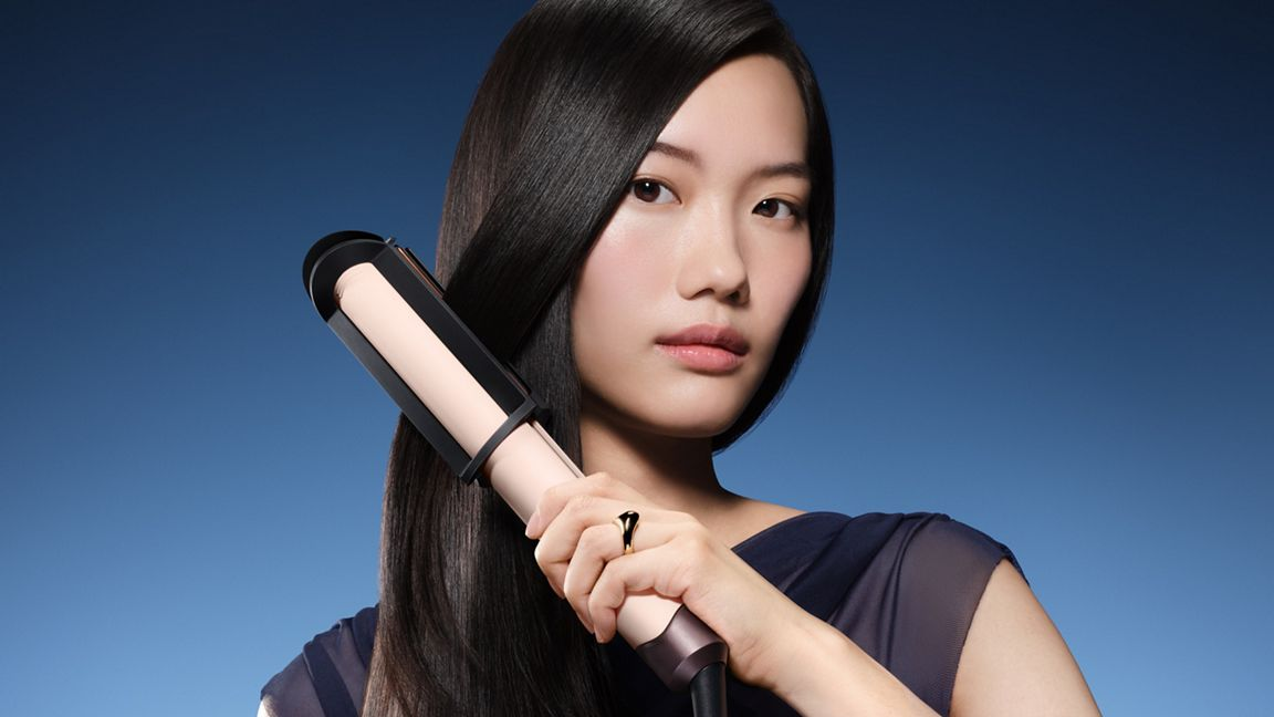 Eine asiatische Frau glättet ihr langes, schwarzes Haar mit einem rosa-schwarzen Glätteisen vor blauem Hintergrund.