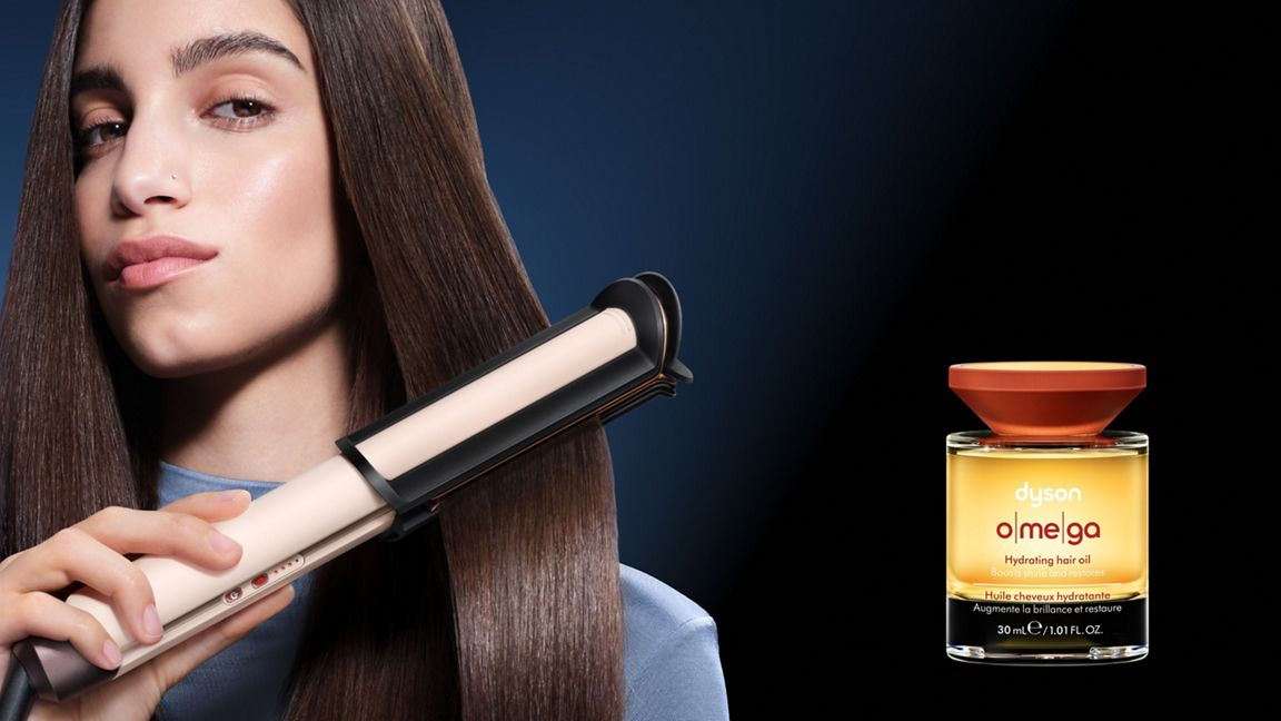 Eine Frau stylt ihr braunes Haar mit einem Glätteisen neben einer Flasche Dyson Haaröl vor dunklem Hintergrund.