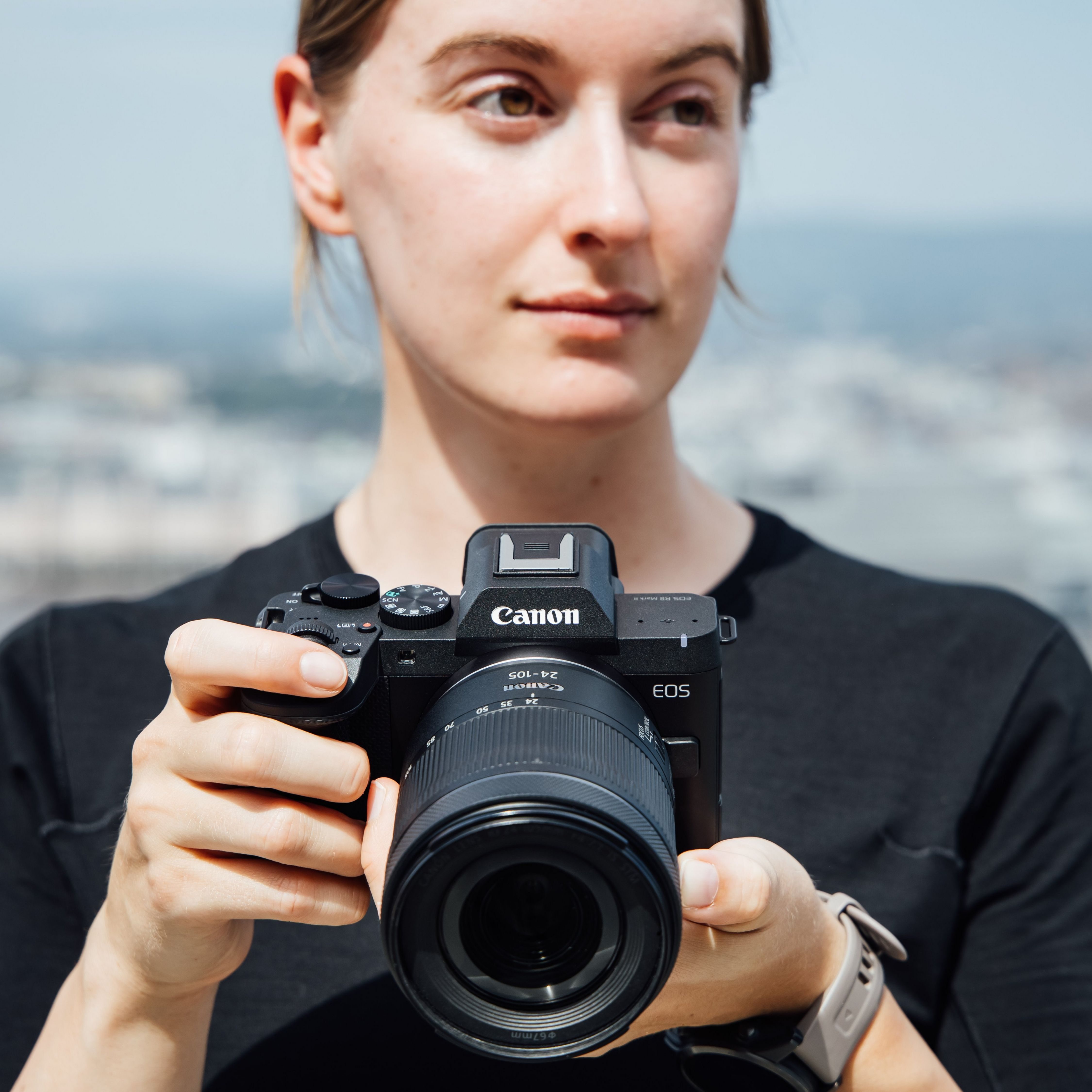 Eine Frau in Schwarz hält eine Canon-Kamera mit Objektiv vor eine verschwommene Stadtlandschaft.