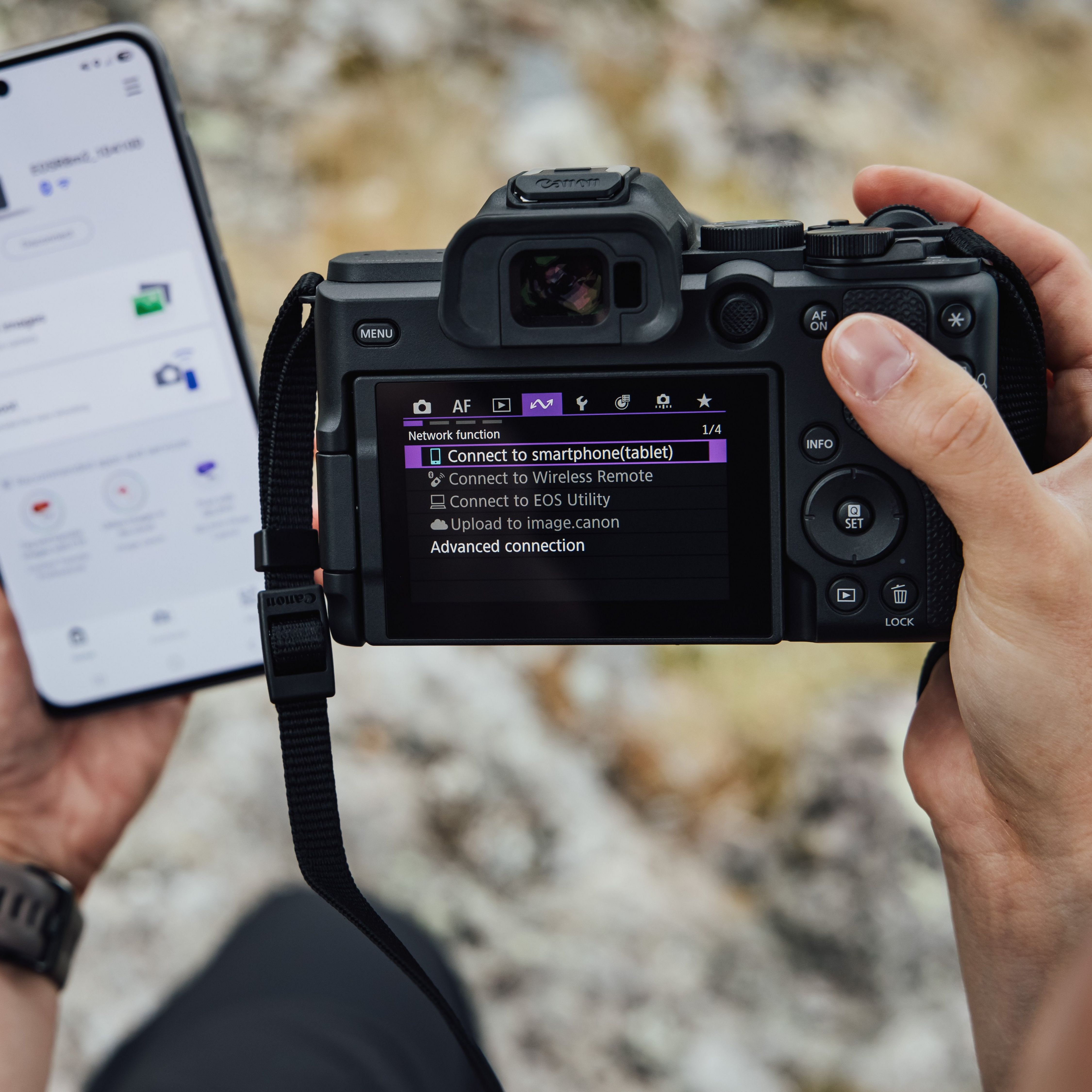 Eine Hand hält eine Canon-Kamera mit Netzwerkeinstellungsmenü auf dem Bildschirm, daneben ein Smartphone.