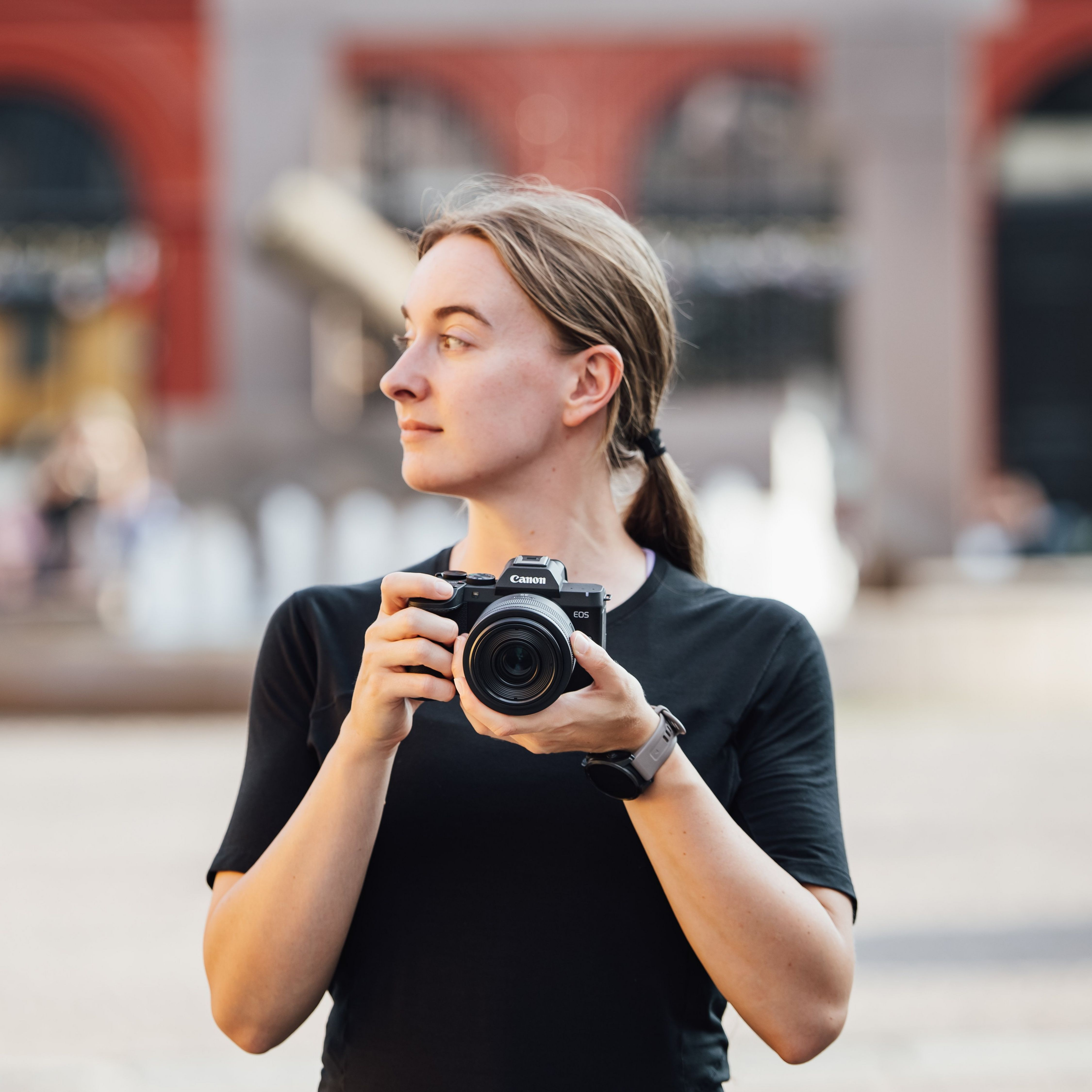 Eine junge Frau in schwarzem Shirt hält eine Canon-Kamera im Freien mit verschwommenem Hintergrund.