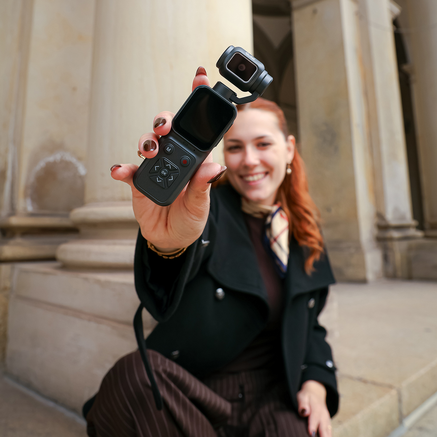 Une femme souriante tient une petite caméra stabilisée face à l'objectif, devant une architecture à colonnes.