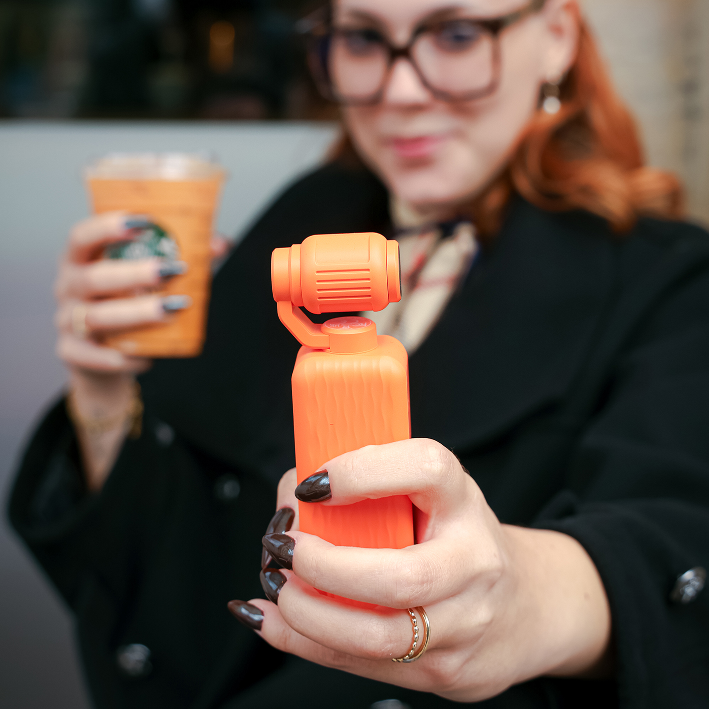 Une femme en manteau noir tient un stabilisateur à main orange et une tasse.