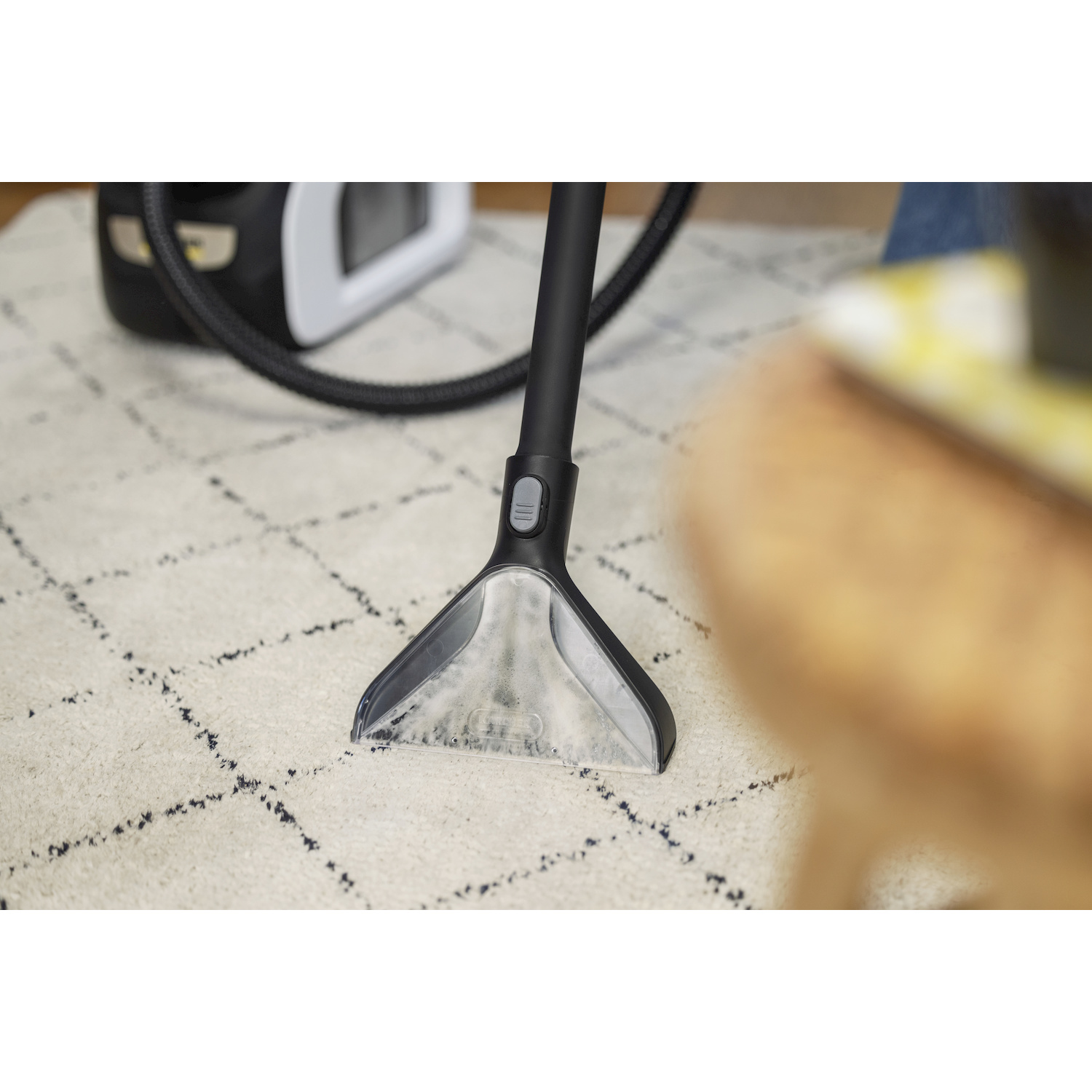 Un pulitore per tappeti aspira su un tappeto bianco a quadri, con l'unità principale sullo sfondo.