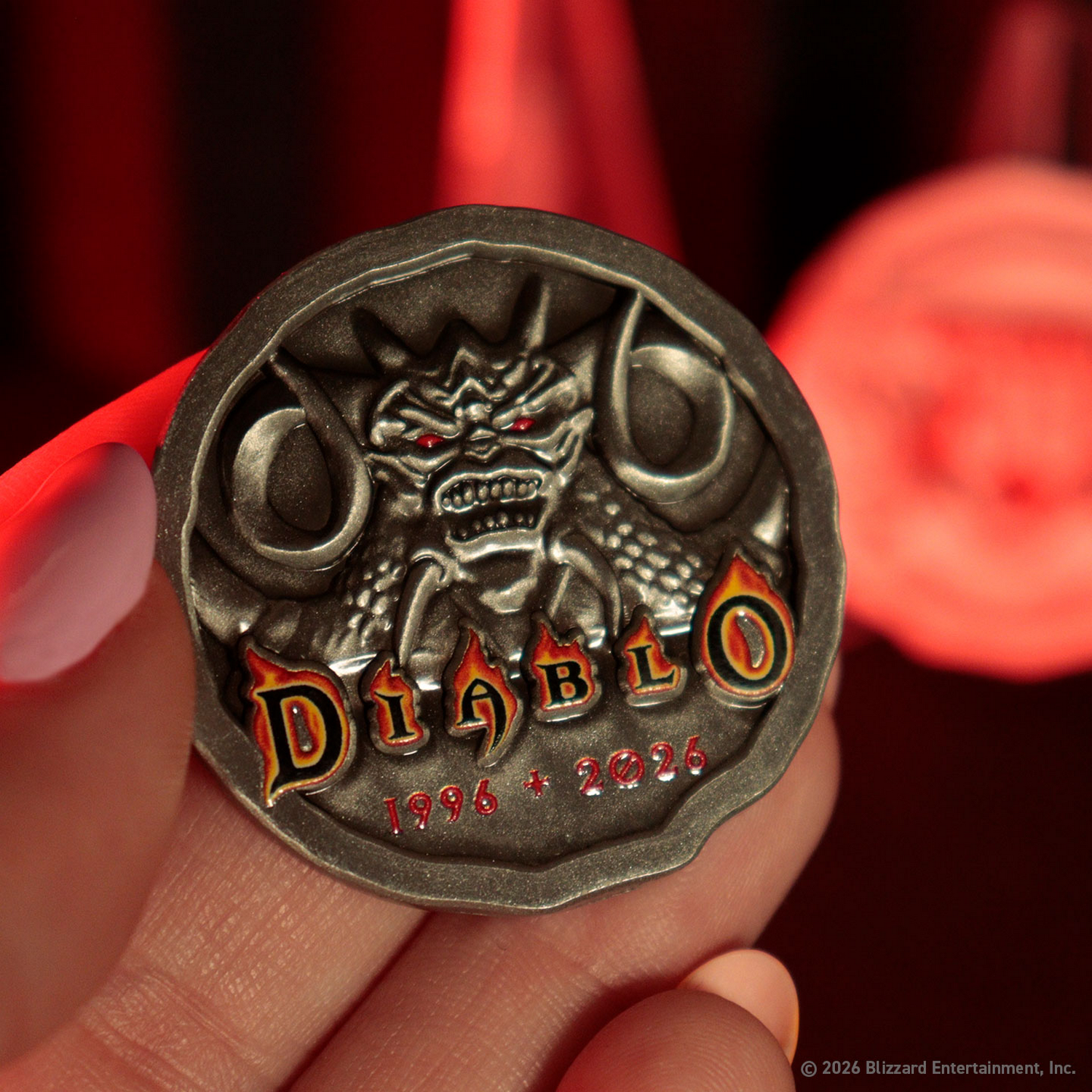 Una mano sostiene una moneda plateada de Diablo con un demonio de ojos rojos y los años 1996 a 2026.