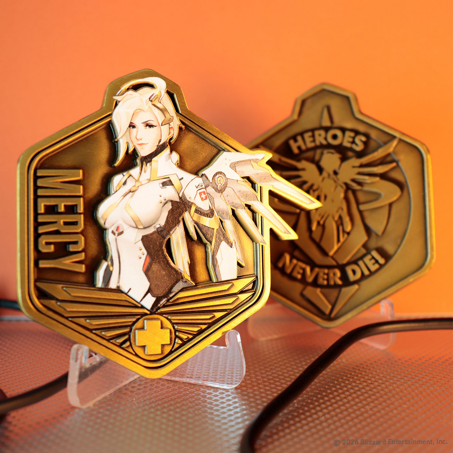 Dos monedas doradas de Mercy de Overwatch en soportes, con Mercy en una y Heroes Never Die en la otra.