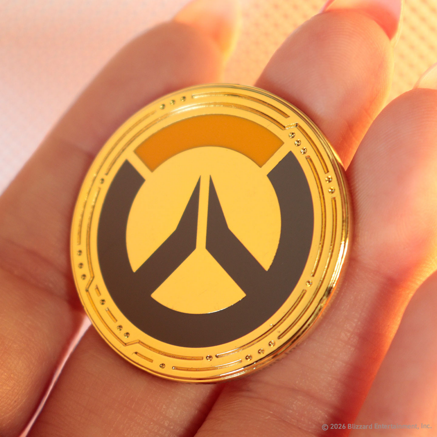Una mano sostiene una moneda de oro con el logotipo de Overwatch en amarillo y negro.