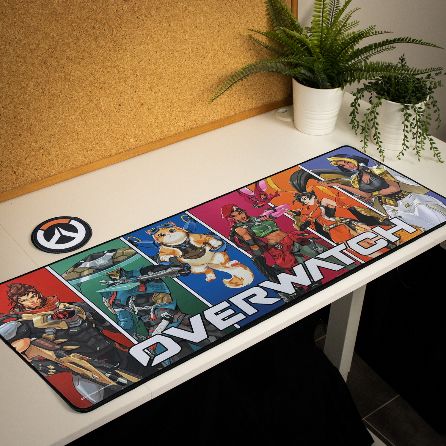 Un tapete de escritorio de Overwatch con arte de personajes en un escritorio blanco, junto a dos plantas y un corcho.