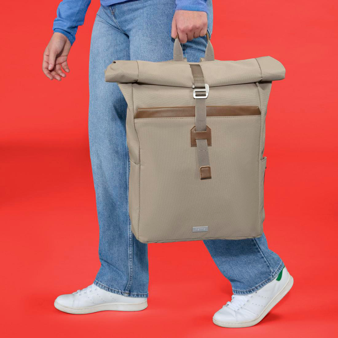 Person in blauen Jeans und weißem Sneaker trägt einen beigen Rucksack vor rotem Hintergrund.