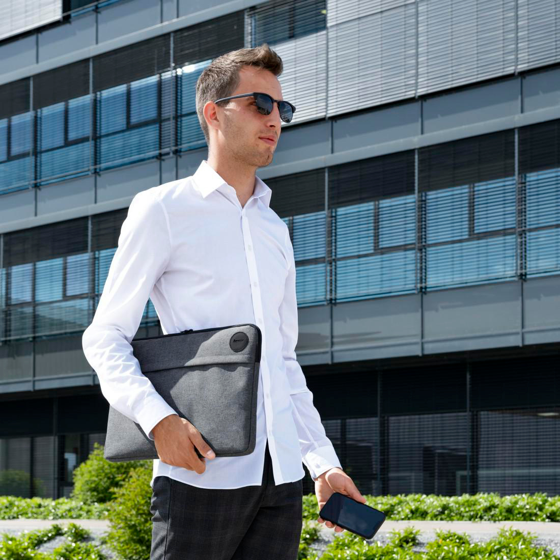 Ein Mann im weißen Hemd und mit Sonnenbrille geht vor einem Bürogebäude und hält eine Laptoptasche und ein Smartphone.