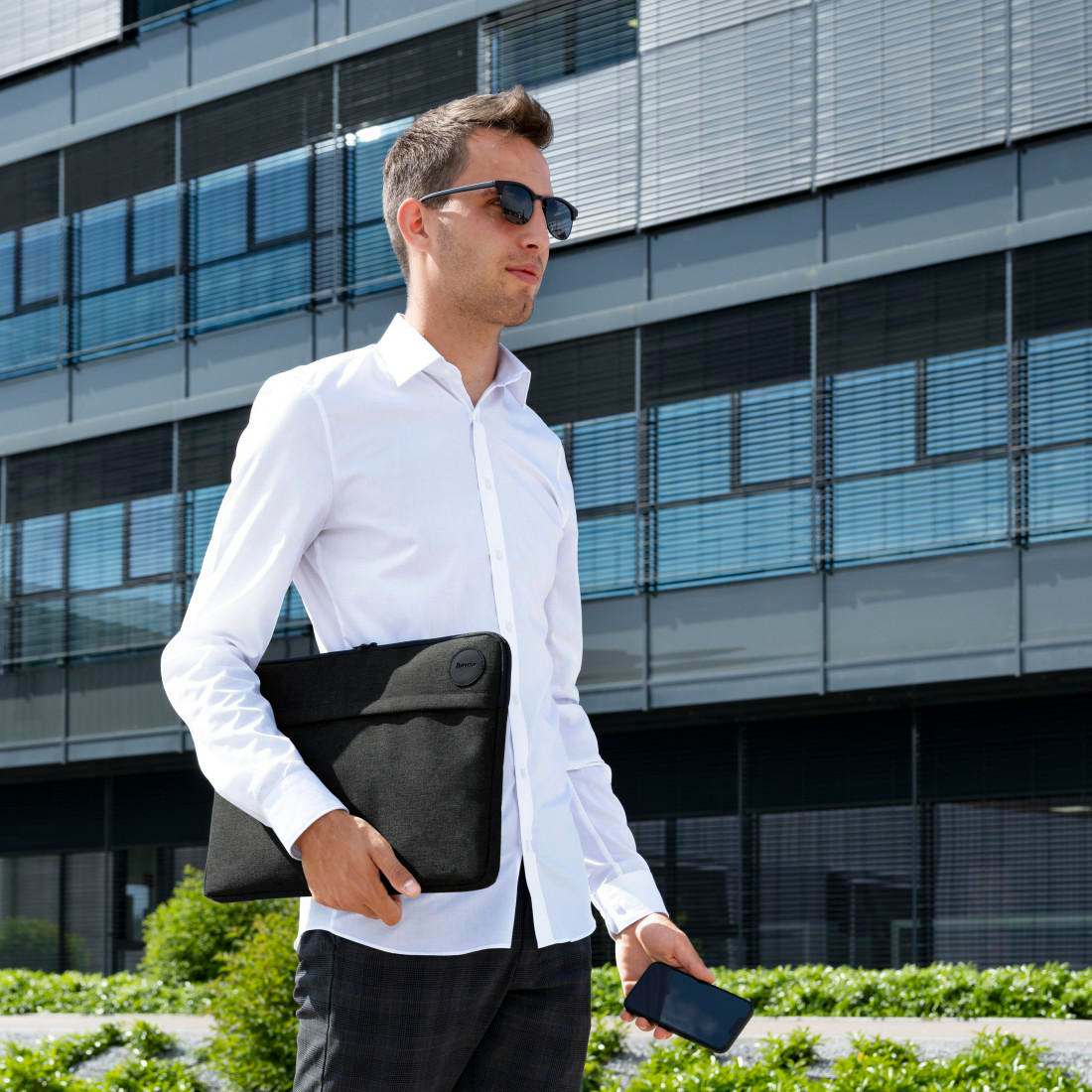 Ein Mann im weißen Hemd und mit Sonnenbrille steht vor einem Bürogebäude und hält eine Laptoptasche.