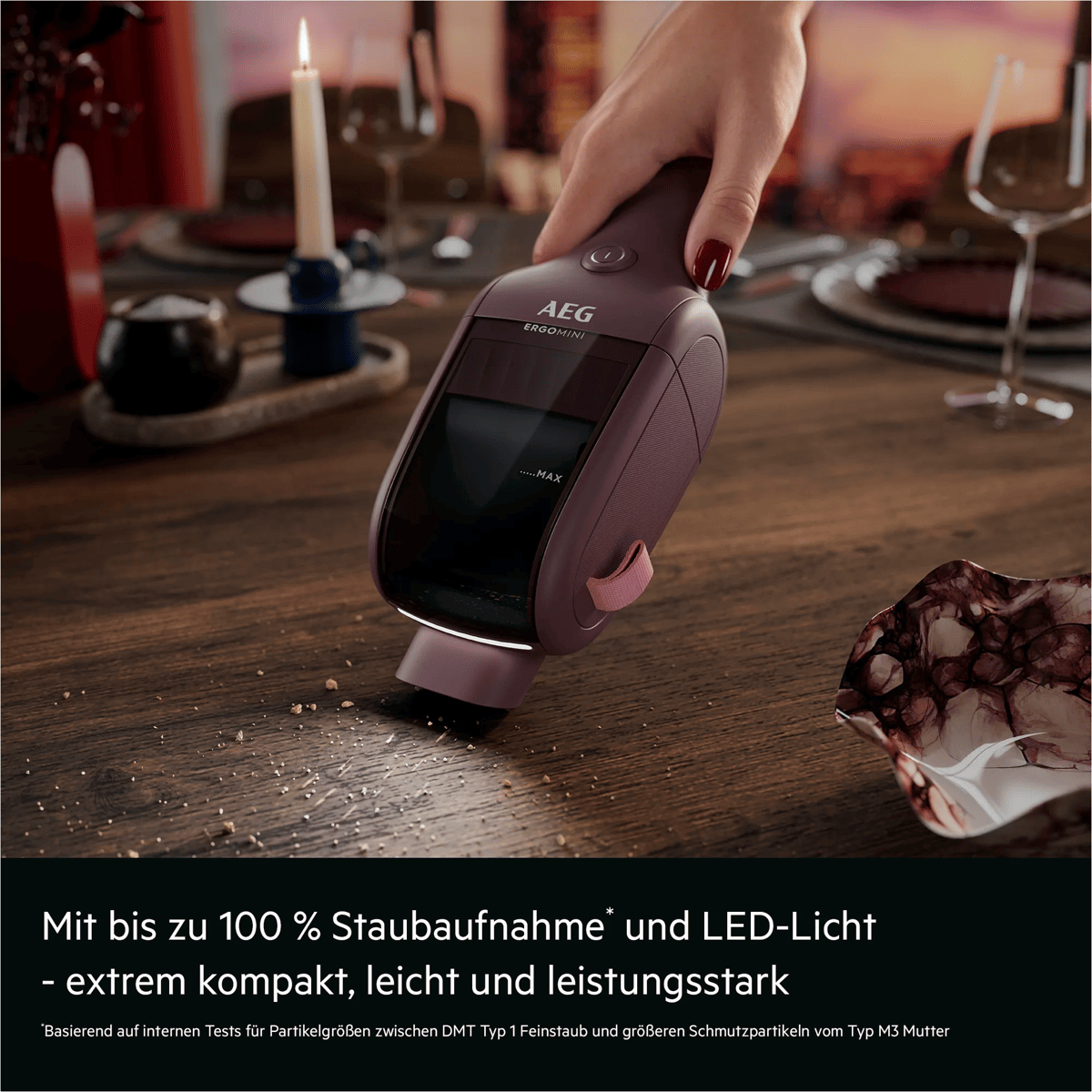 Eine Hand hält einen violetten AEG Handstaubsauger, der Krümel von einem Holztisch saugt.