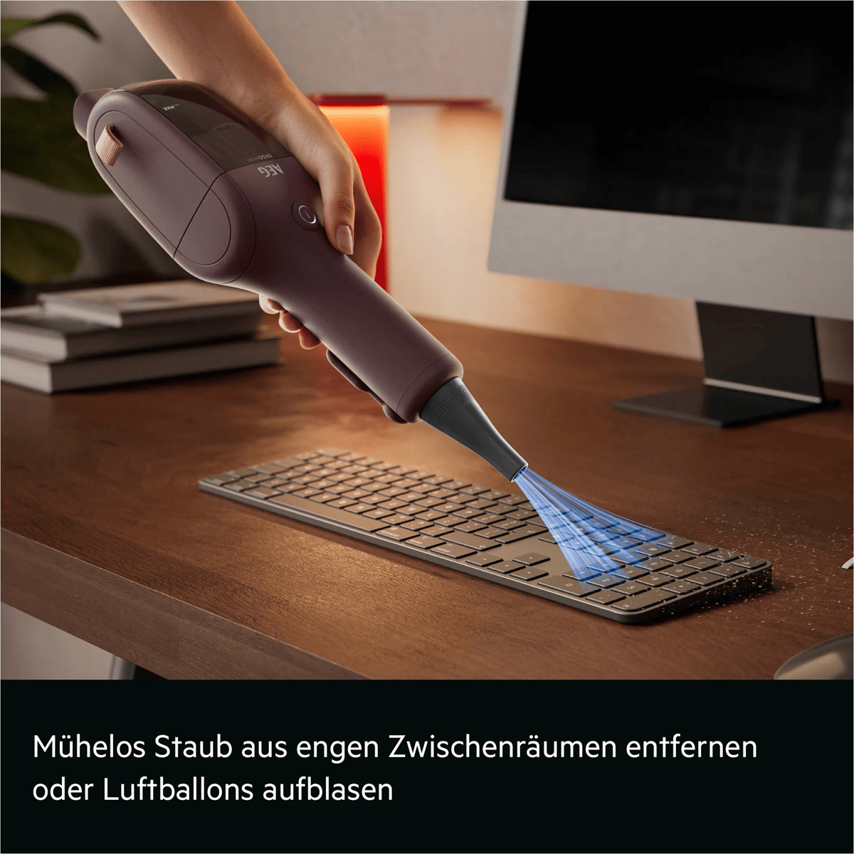 Ein Handstaubsauger bläst mit einem feinen Strahl Staub von einer Computertastatur auf einem Holzschreibtisch.