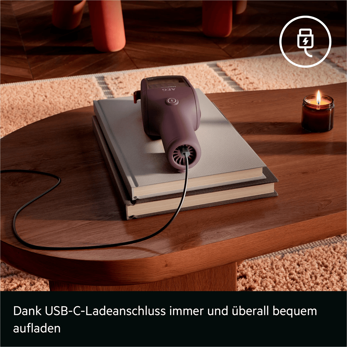 Ein violetter Handstaubsauger lädt auf Büchern auf einem Holztisch, neben einer Kerze und einem USB-Symbol.