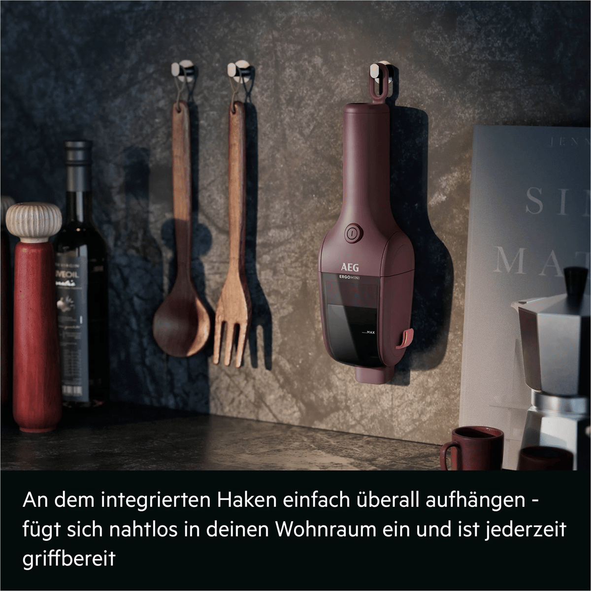 Ein maronifarbener AEG Handstaubsauger hängt an einer dunklen Küchenwand neben Holzlöffeln und einer Kaffeemaschine.