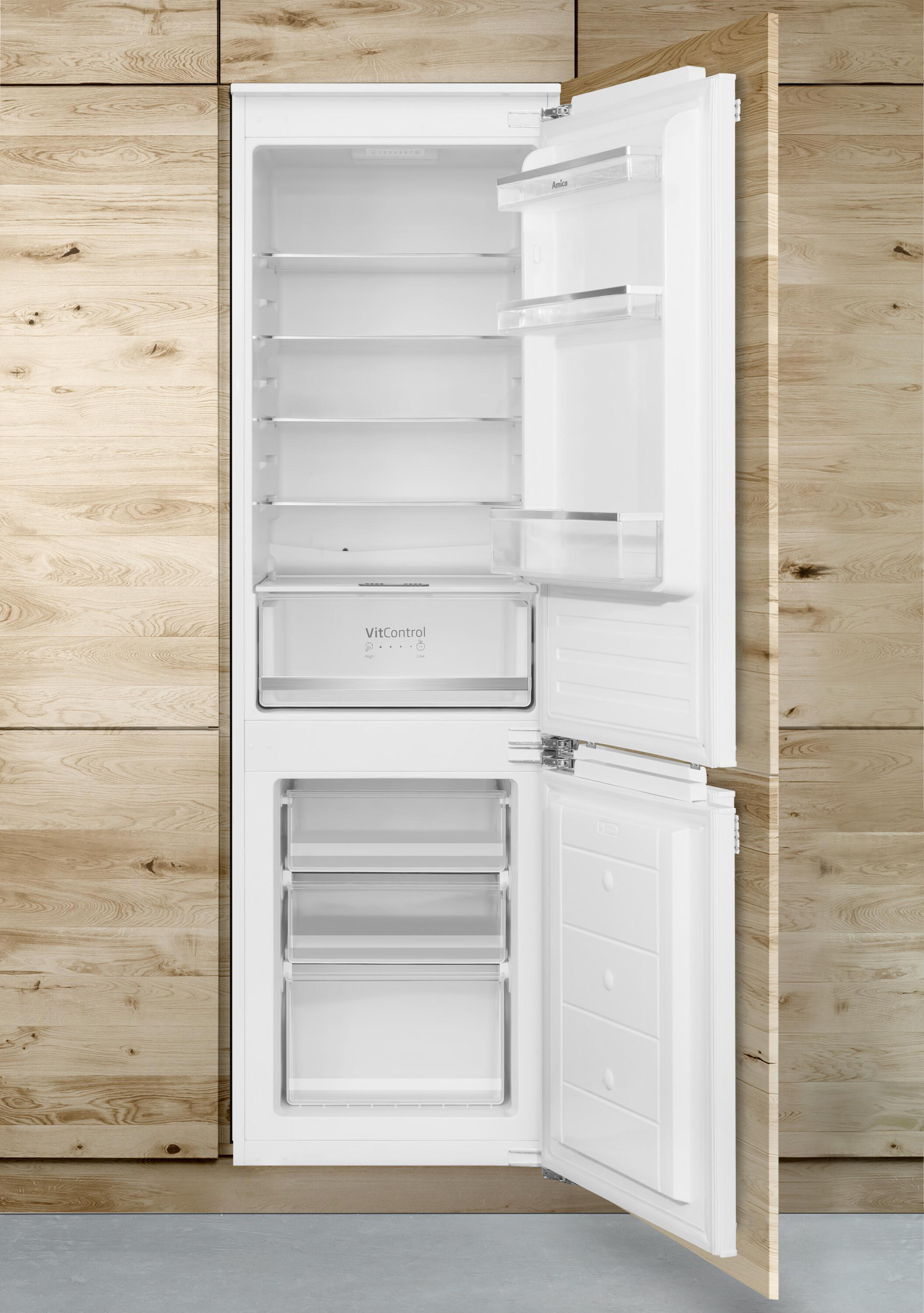 Ein weißer, offener Einbaukühlschrank mit Glasböden und Schubladen vor einer hellen Holzwand.