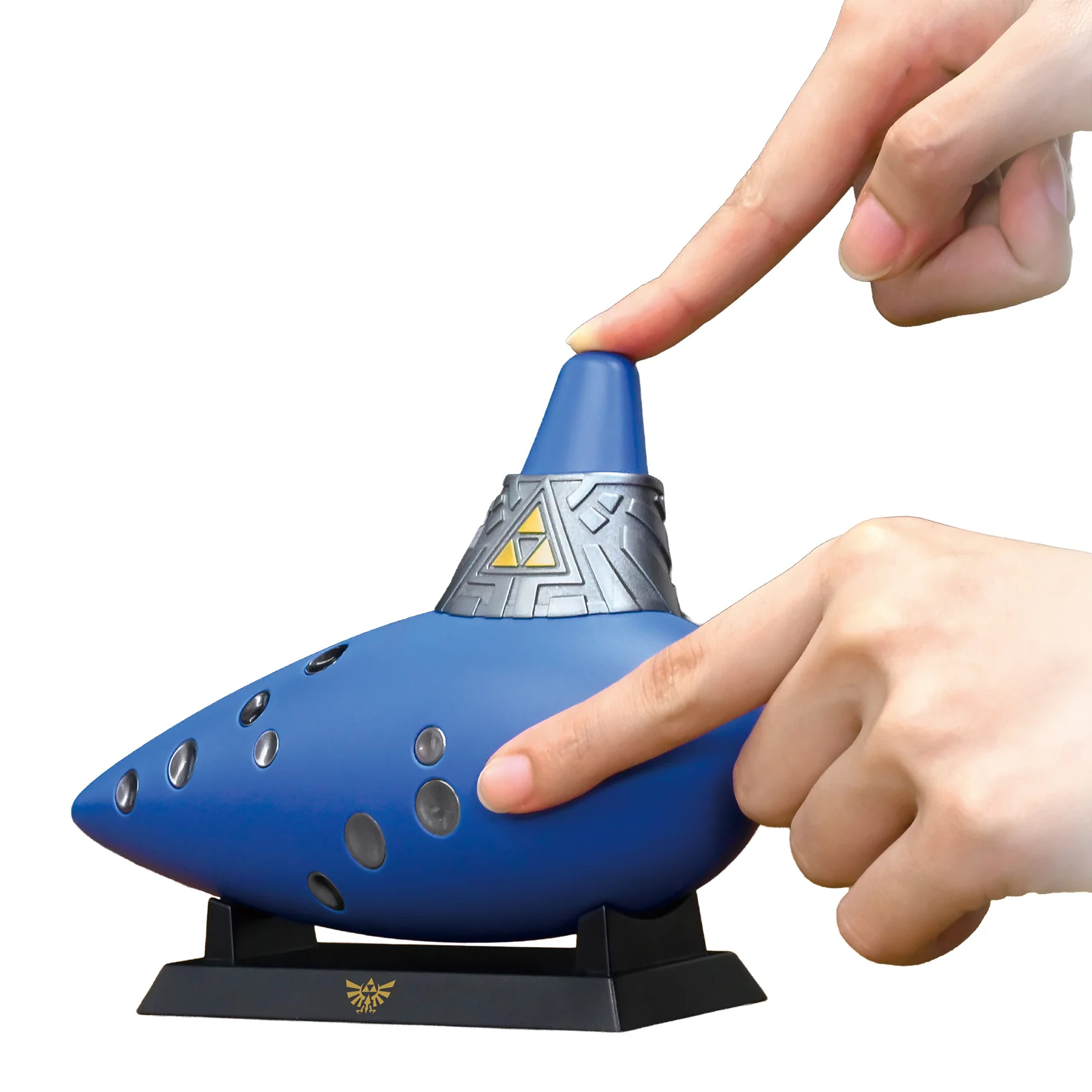 Manos tocando una ocarina azul con símbolo Trifuerza dorado en un soporte.