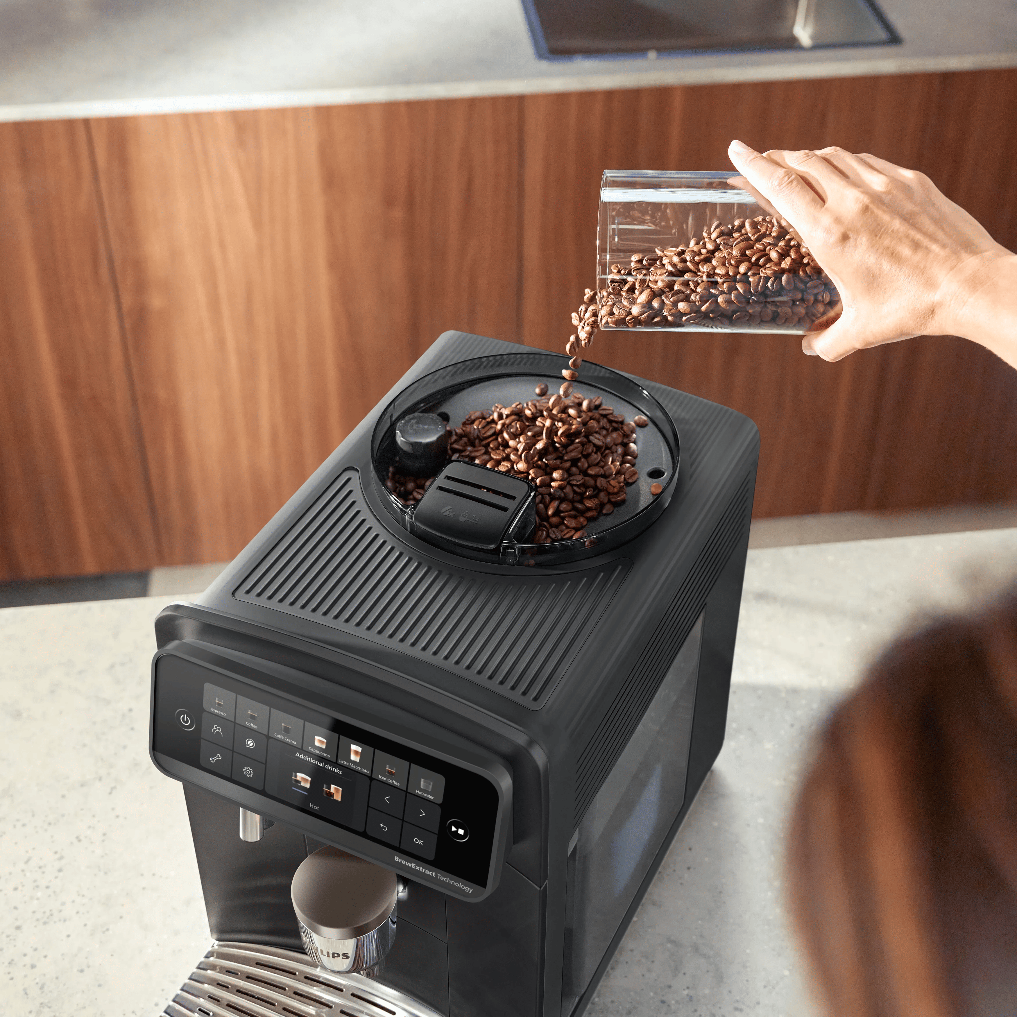 Eine Hand schüttet Kaffeebohnen aus einem Behälter in den Bohnenbehälter einer modernen schwarzen Kaffeemaschine.