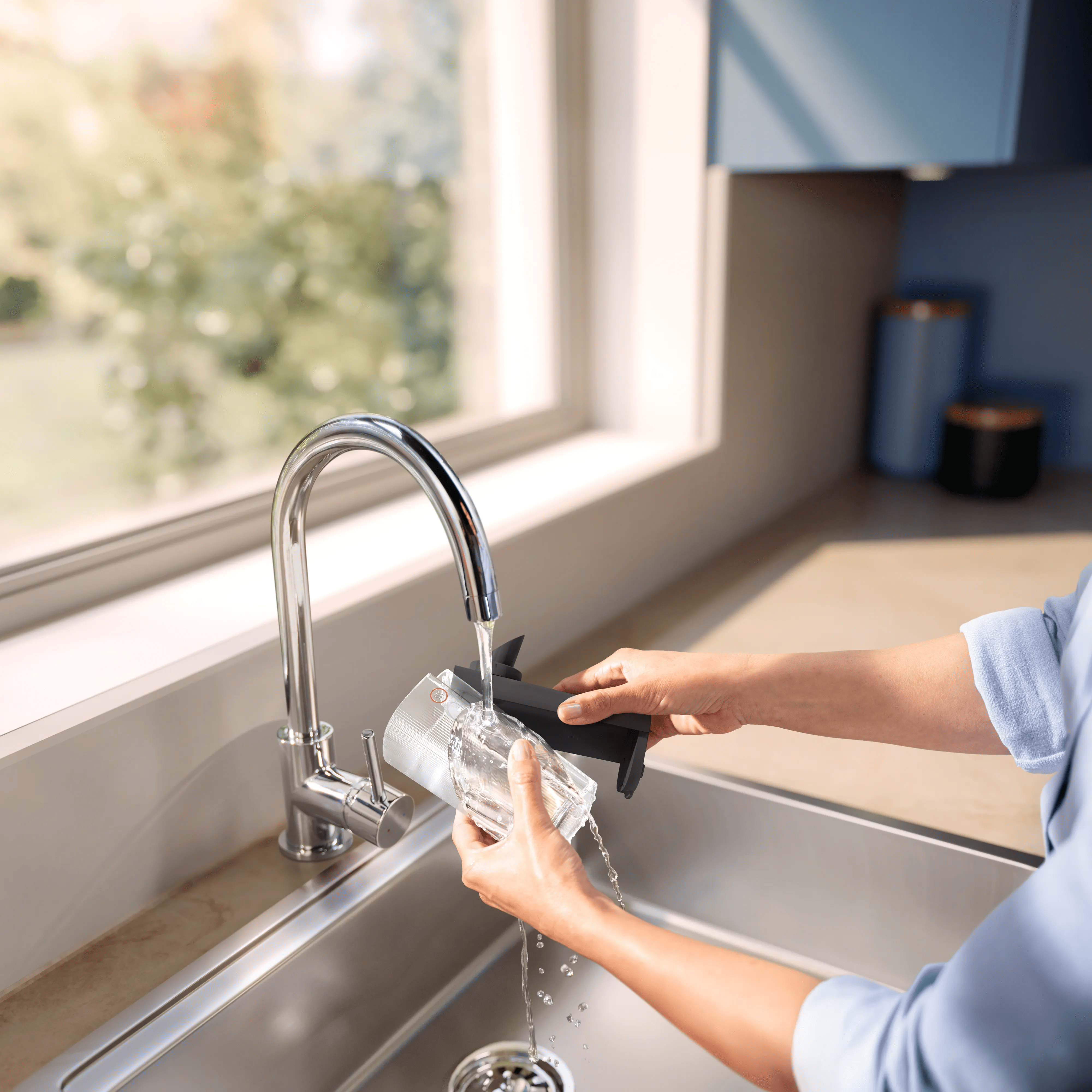 Person wäscht einen kleinen Behälter unter dem Wasserhahn in einer hellen Küche am Fenster.