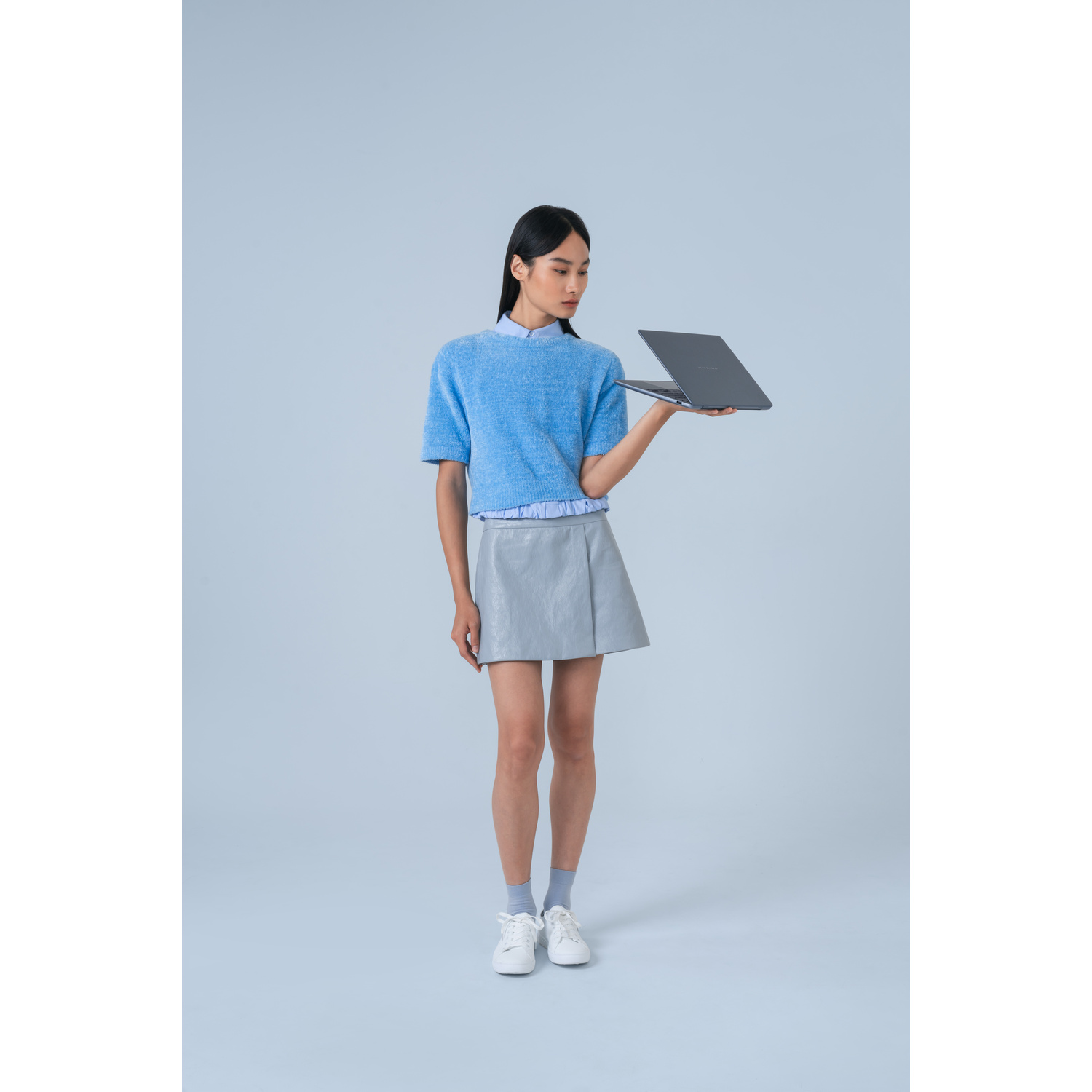Una donna con maglione blu e gonna grigia tiene un laptop aperto su sfondo blu.