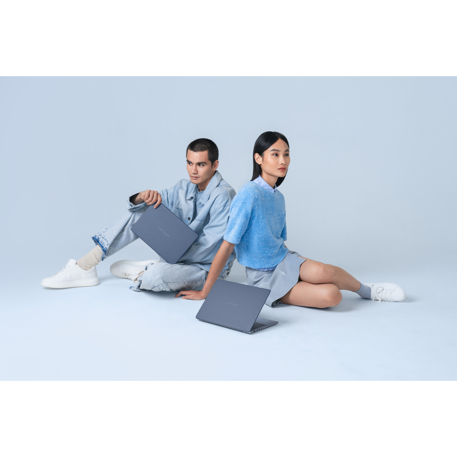 Un giovane e una donna in abiti blu siedono sul pavimento, ciascuno con un computer portatile blu in mano.