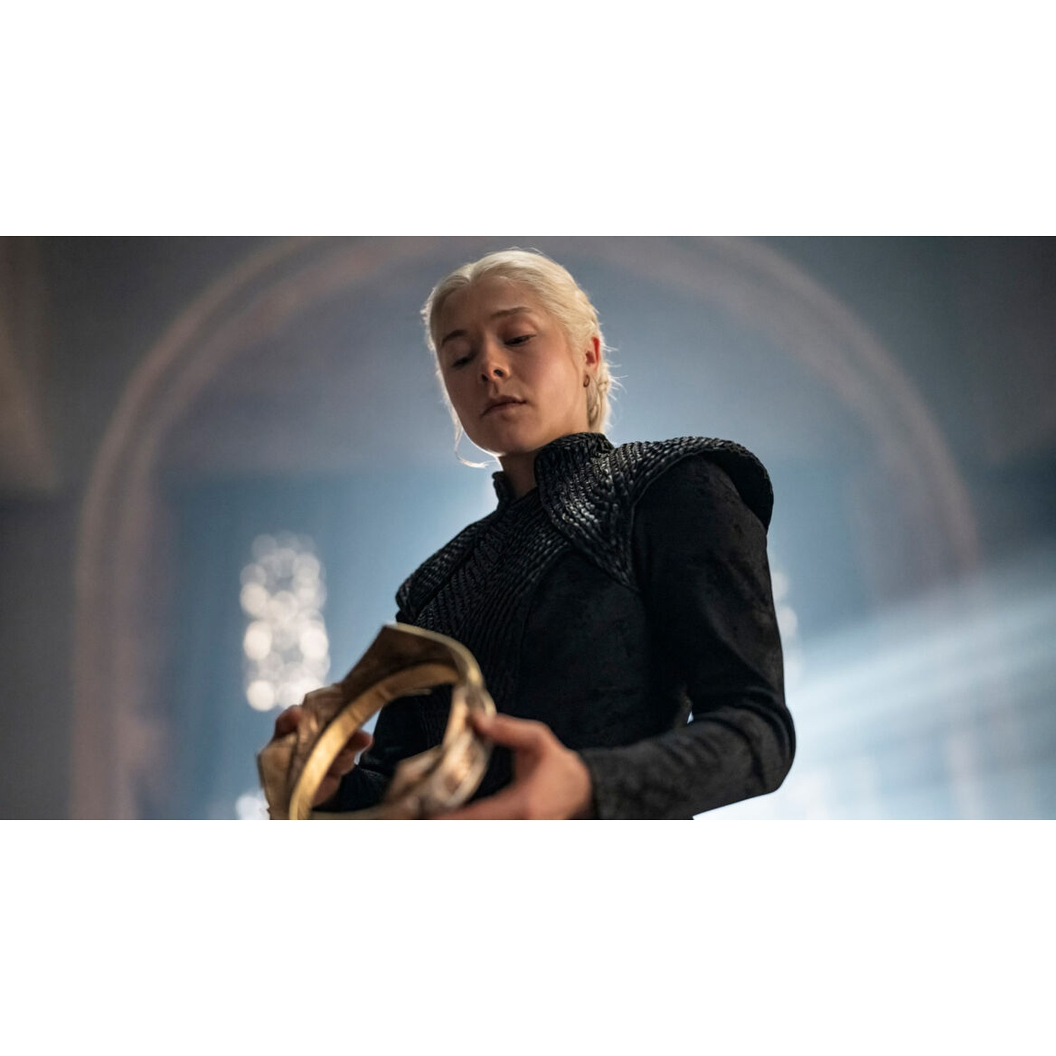 A blonde woman in black clothing holds a golden crown in her hands.