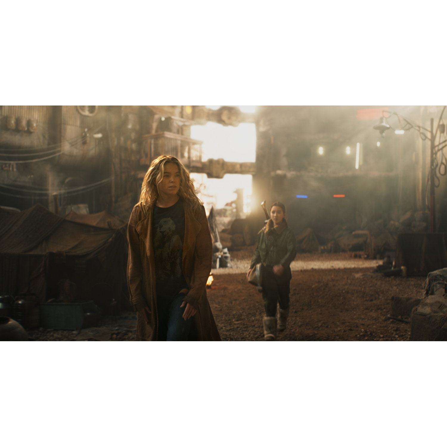 A blonde woman in a brown coat walks in a dusty industrial setting, followed by a young girl.