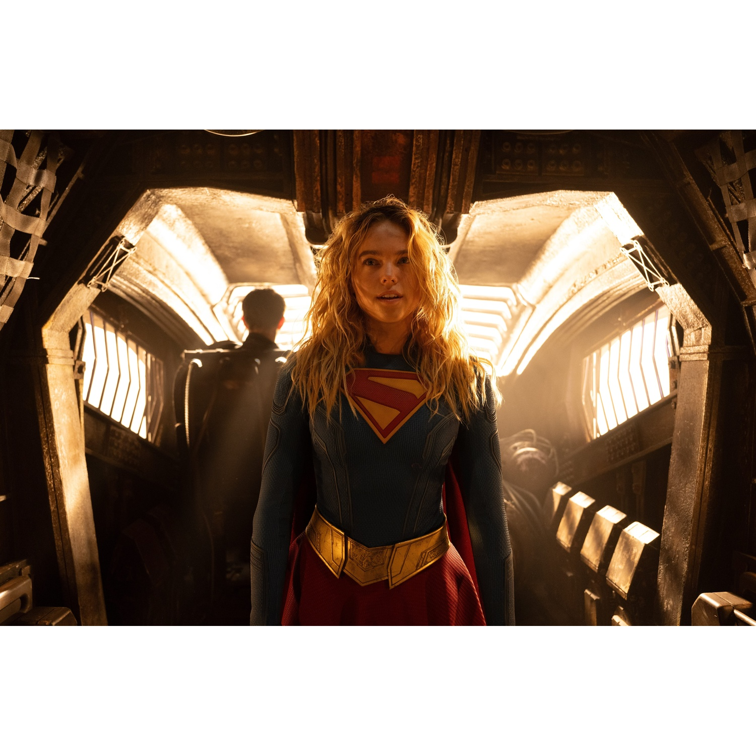 Supergirl with blonde hair and a blue and red costume stands in a futuristic tunnel.