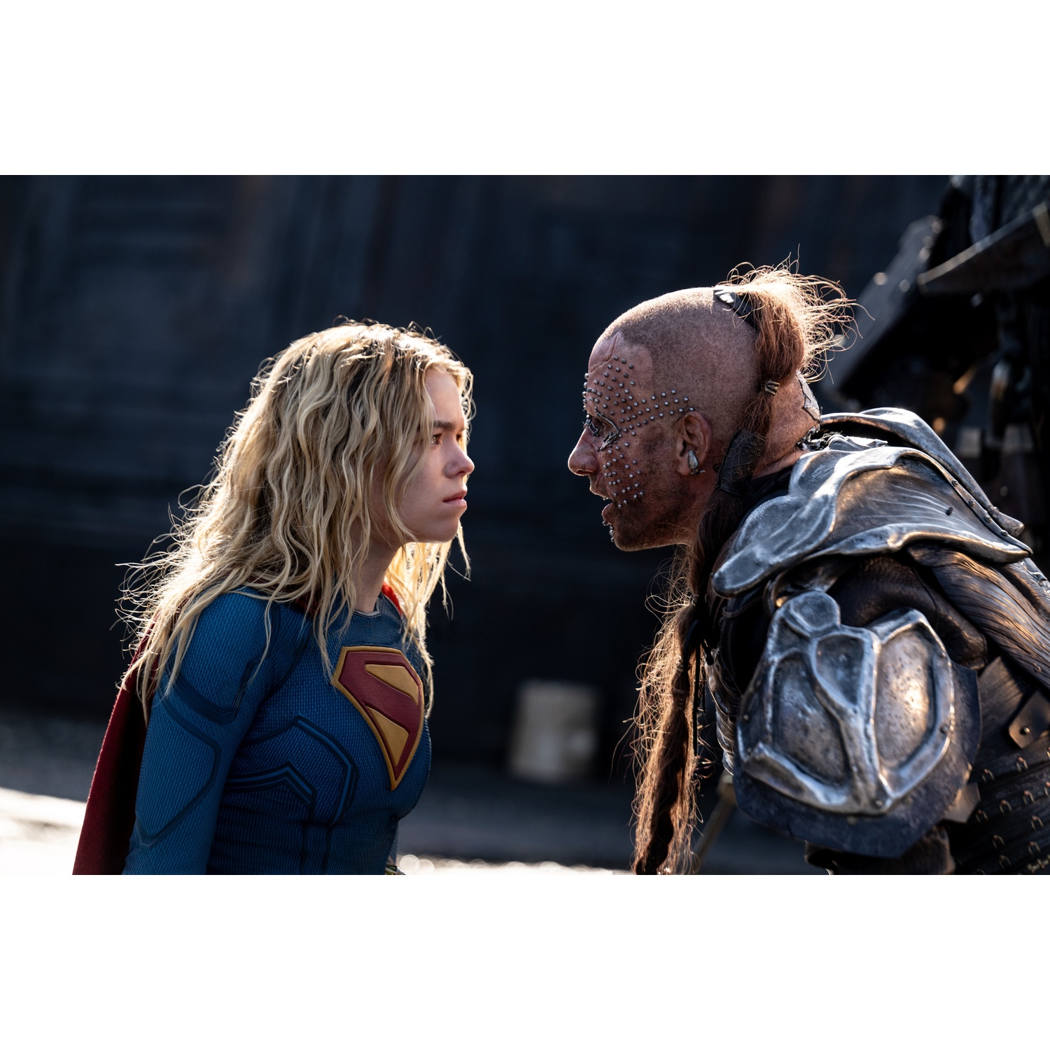 Supergirl with blonde hair and blue suit stares intensely face-to-face with a heavily armored, scarred warrior.