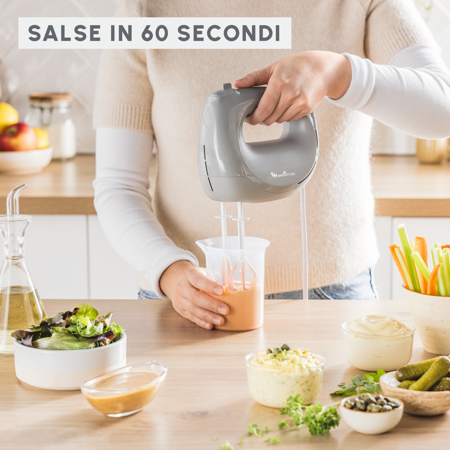 Una persona usa un mixer a mano grigio per frullare la salsa in un contenitore con salse e verdure.