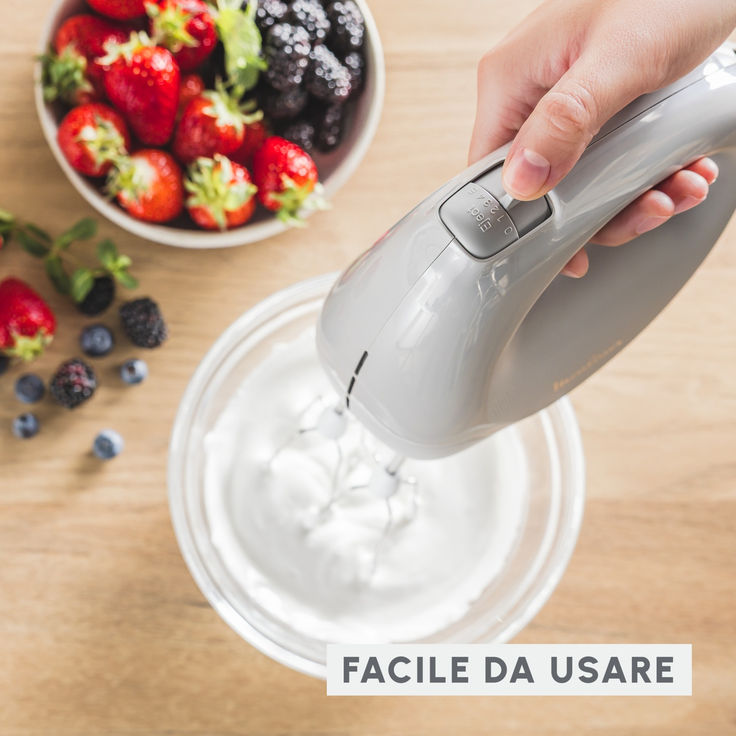Una mano tiene un mixer grigio su una ciotola di crema, con bacche e frutta su un tavolo di legno.