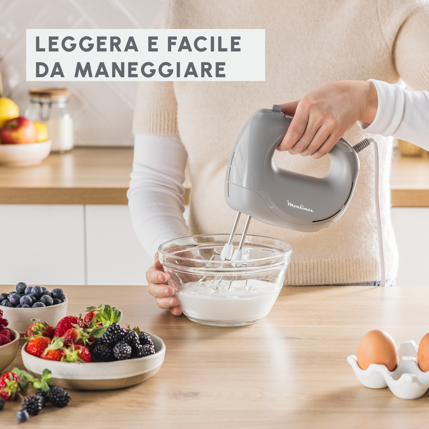 Una donna monta la panna in una ciotola di vetro con un frullatore grigio; frutta fresca e uova sul tavolo.