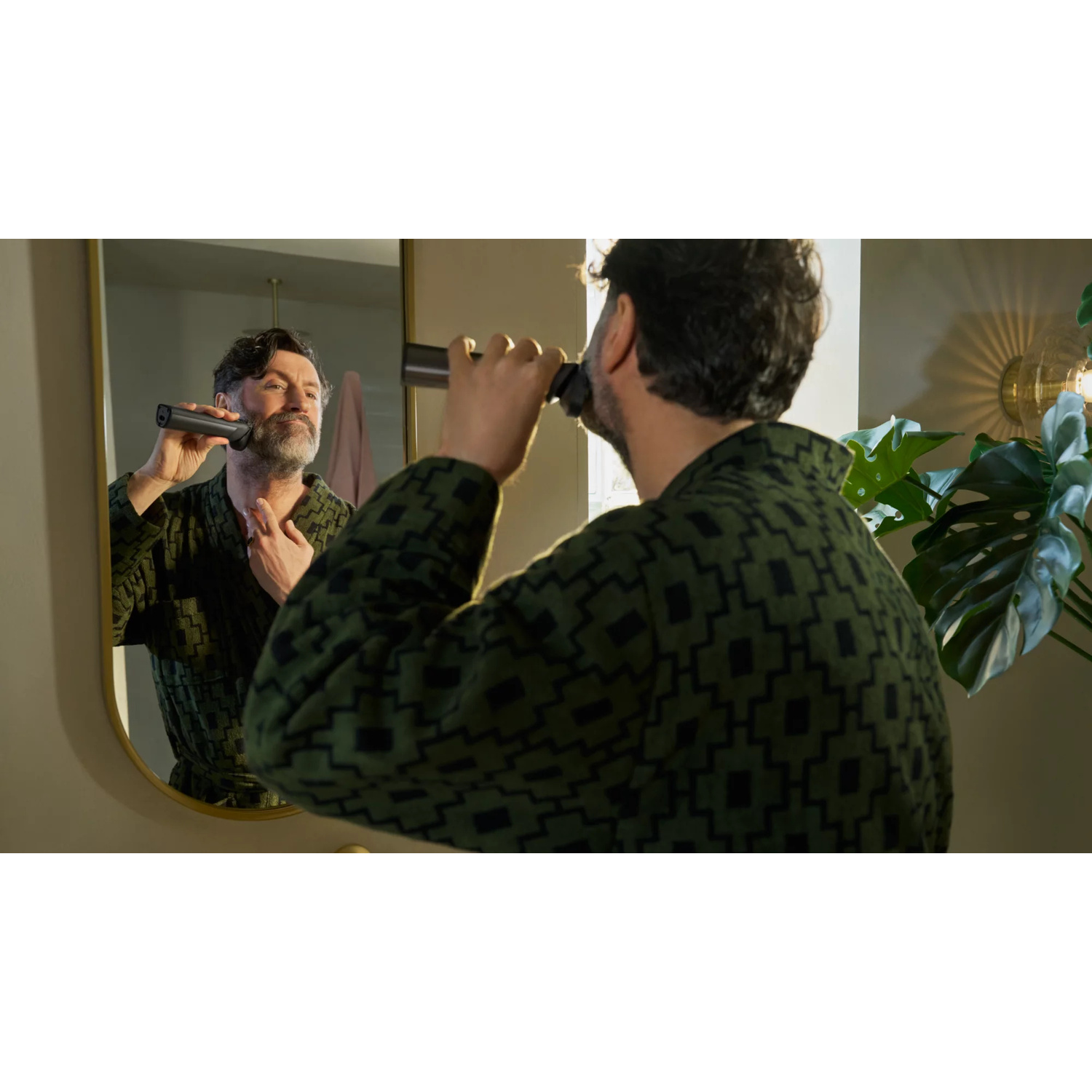 Un uomo in vestaglia verde si sbarba davanti a uno specchio con un rasoio nero.