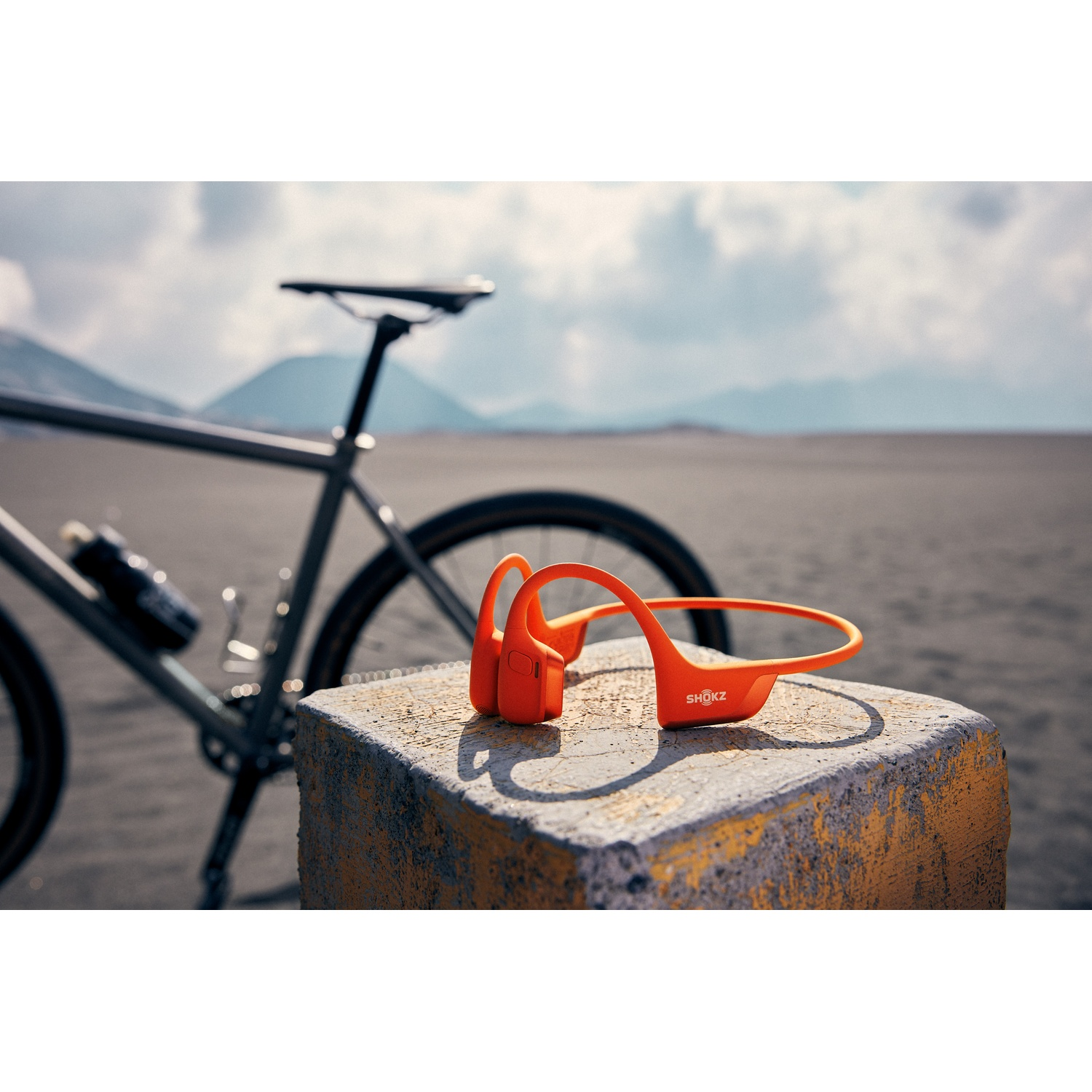 Écouteurs Shokz orange vif sur un bloc de béton avec un vélo et un paysage brumeux en arrière-plan.