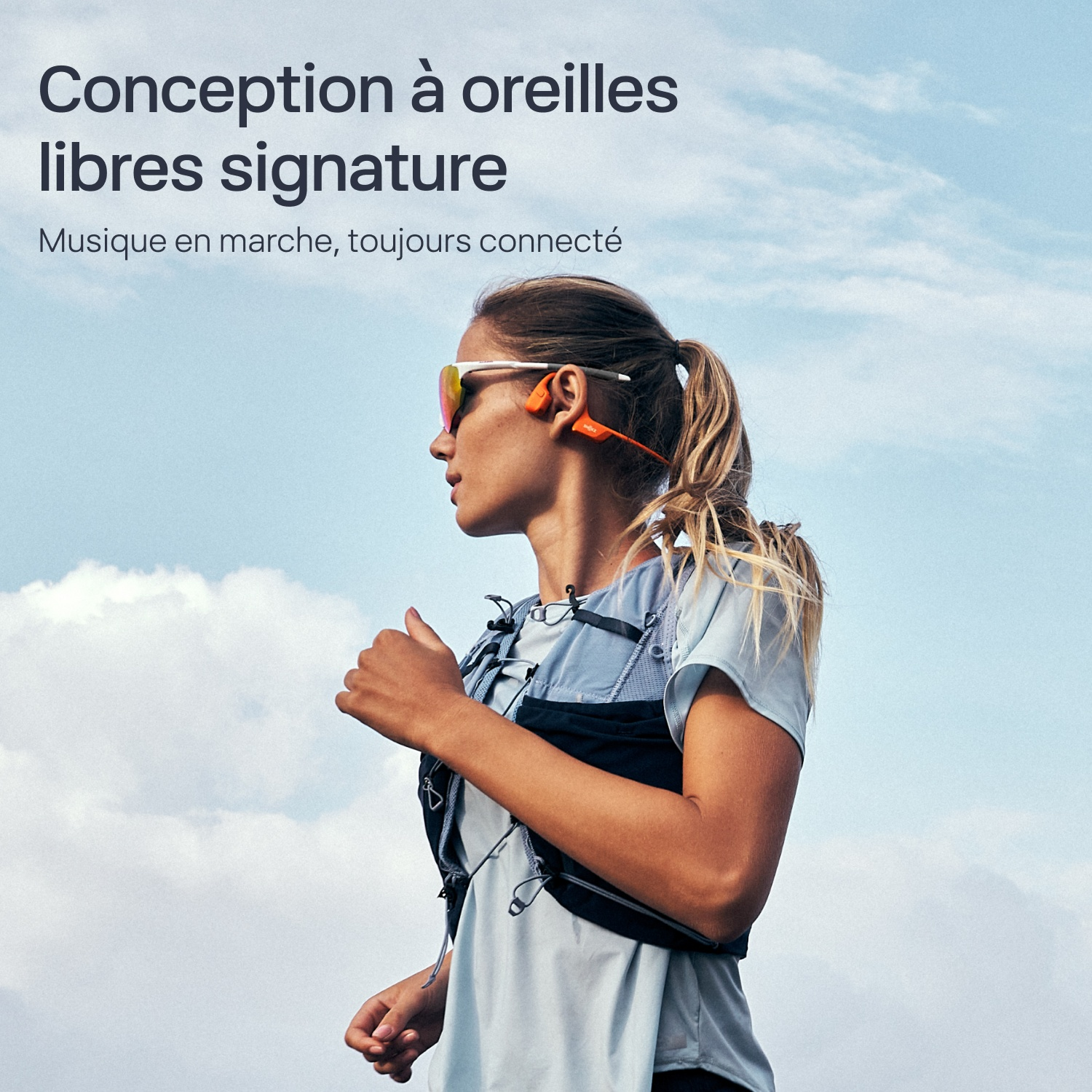 Une femme court en plein air avec des écouteurs à conduction osseuse orange sous un ciel nuageux.