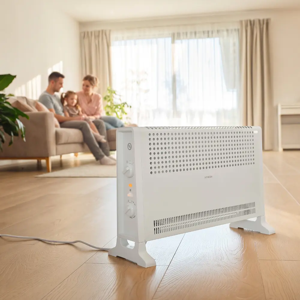 Egy fehér elektromos fűtőtest áll a fa padlón egy világos nappaliban, a háttérben egy kanapén ülő családdal.