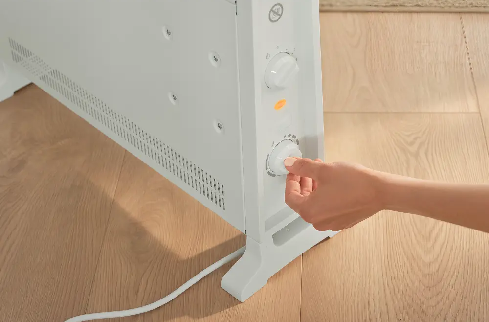Egy kéz állítja be a termosztát gombját egy fehér konvektoron, amely fa padlón áll.