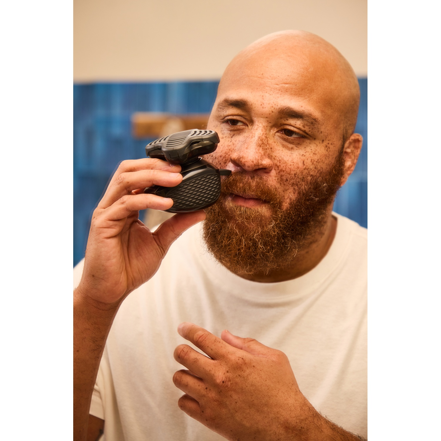 Een kale man met een baard scheert zijn gezicht met een handdrand elektrisch scheerapparat voor een blauwe muur.