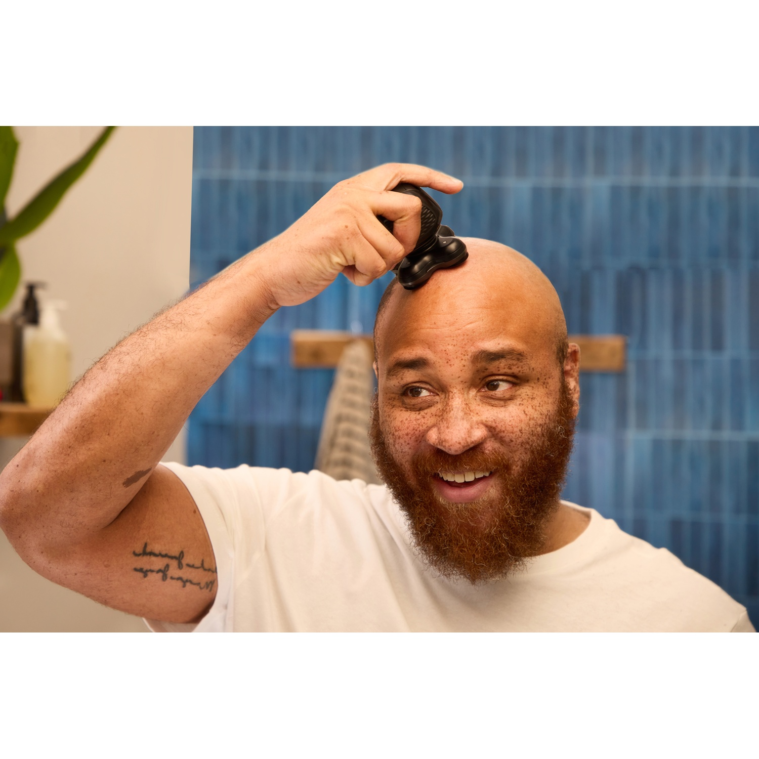 Een lachende man met een baard scheert zijn hoofd met een elektrisch scheerapparaat voor blauwe tegels.