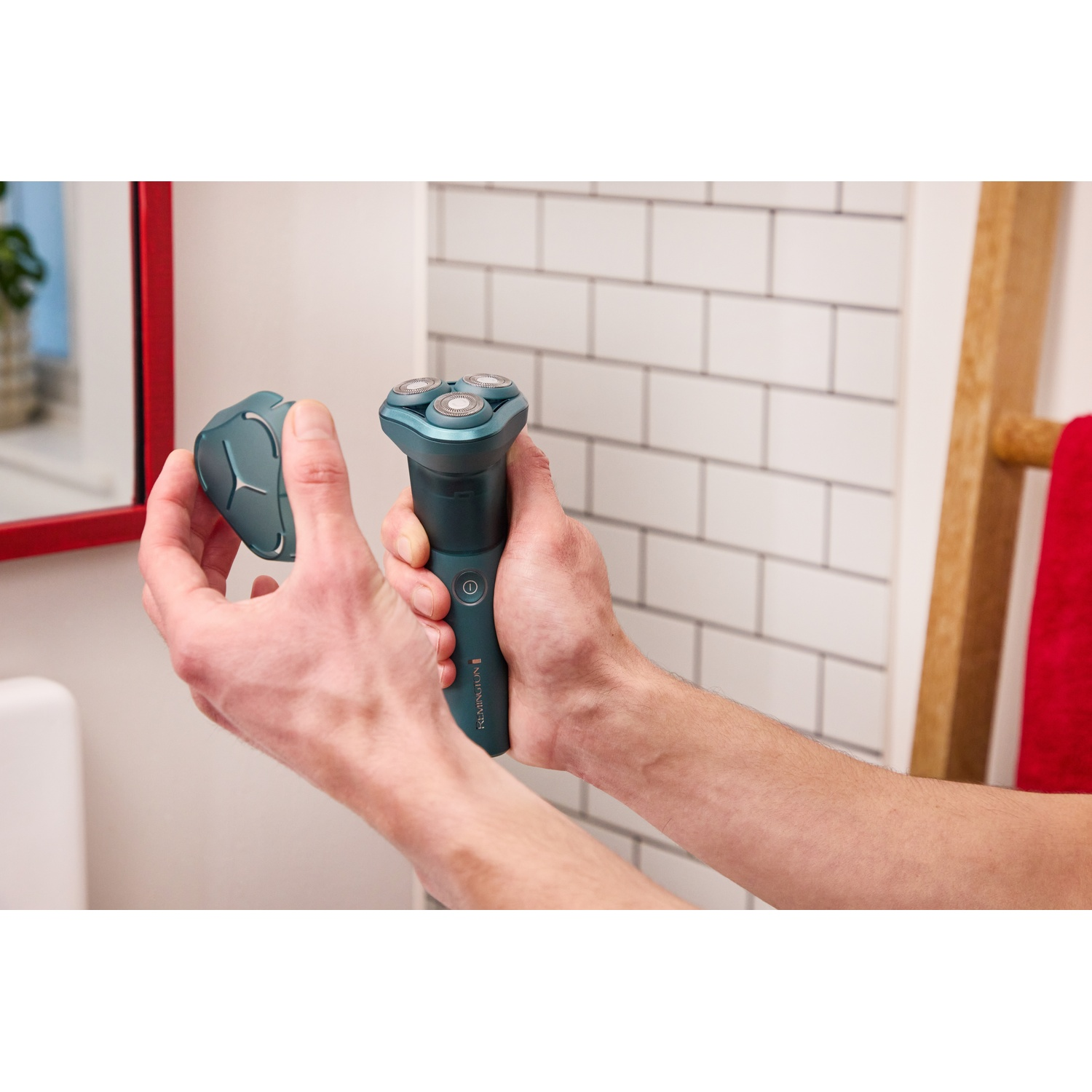Iemand houdt een groenblauw elektrisch scheerapperaat en dop vast voor een witte tegelmuur.