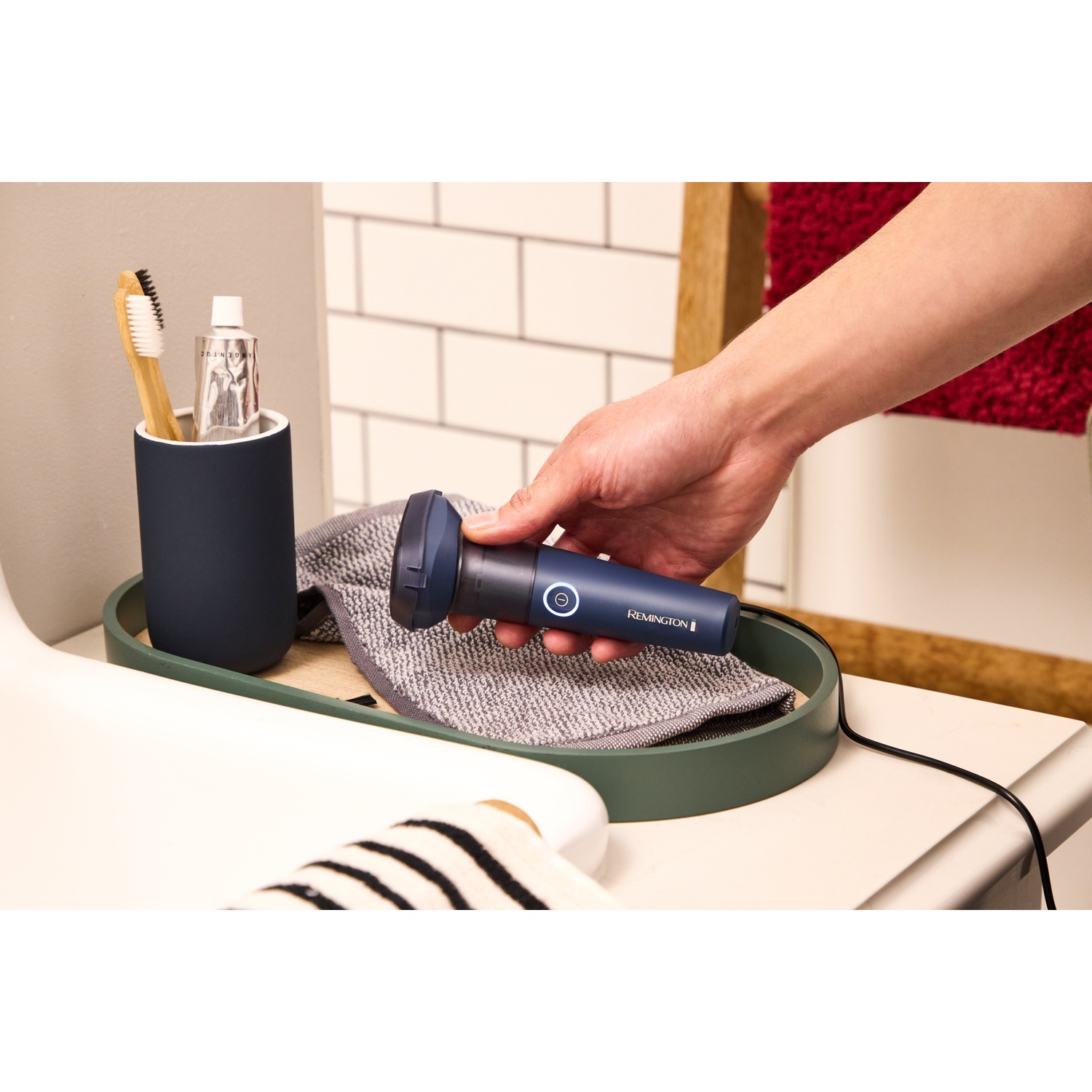 Een hand houdt een blauw Remington scheerapparaat vast in een badkamer naast een tandenborstelhouder en handdoek.