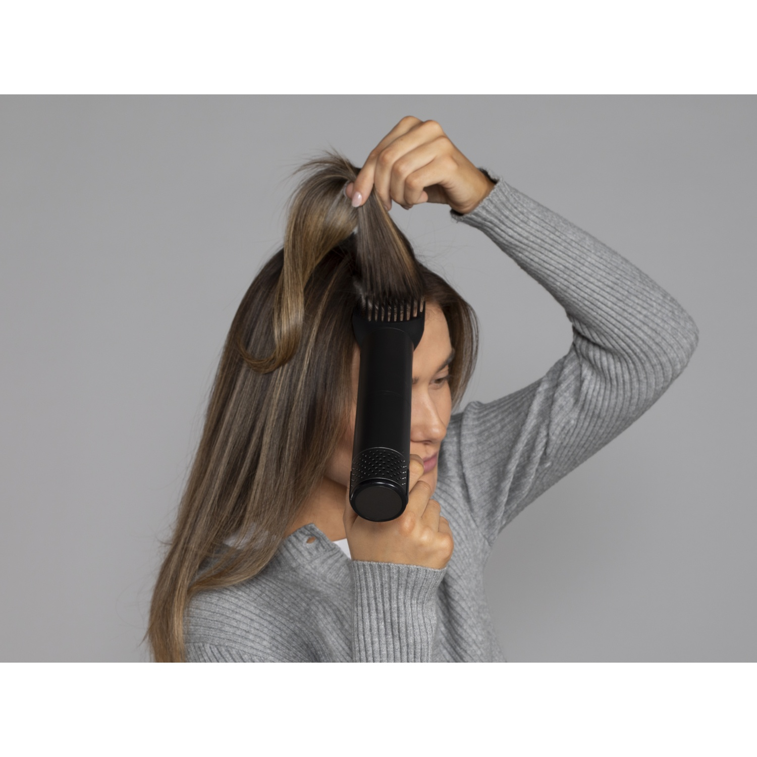 Een vrouw in een grijze trui gebruikt een zwarte haarborstel op haar lange blonde haar tegen een egale achtergrond.