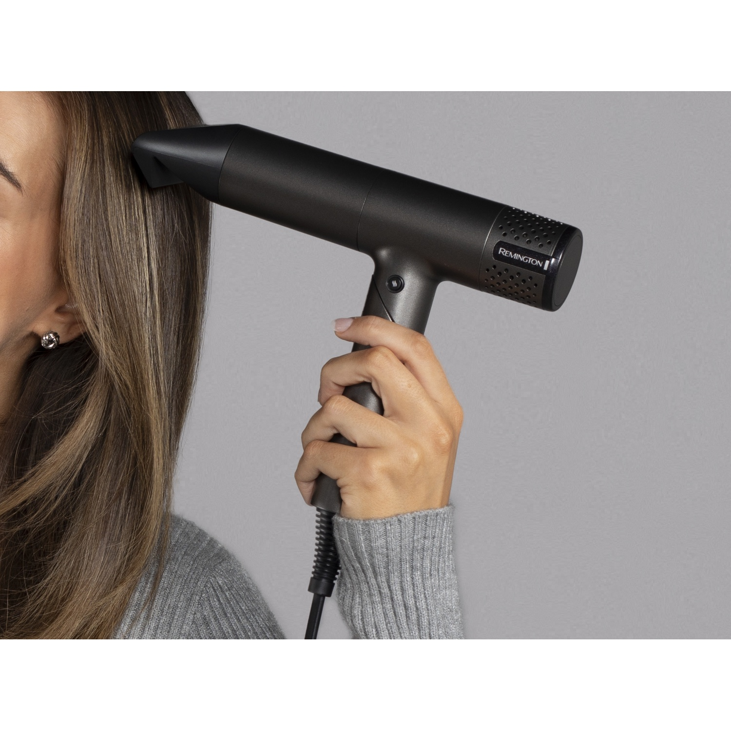 Een vrouw in een grijze trui houdt een zwarte Remington-föhn bij haar haar.