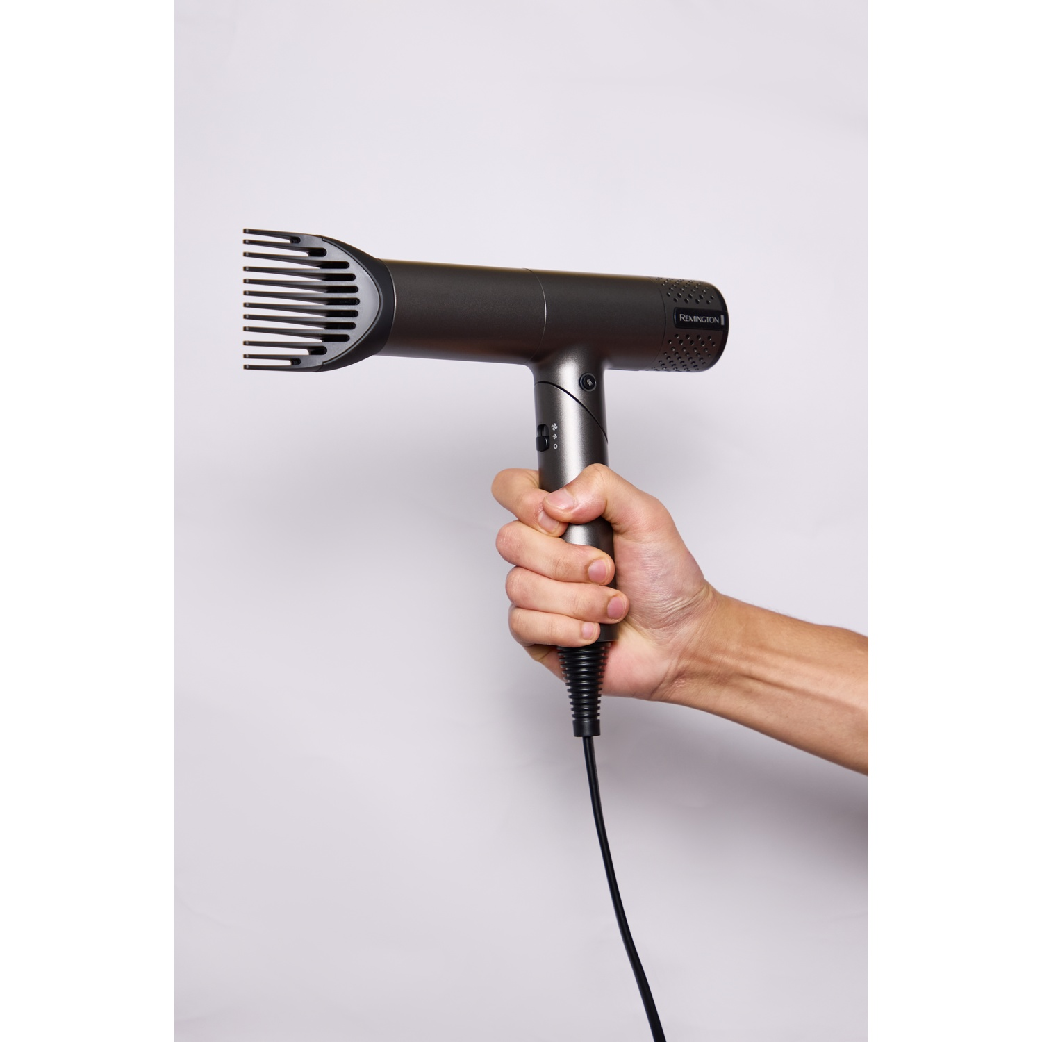 Een hand houdt een grijze Remington haardroger met kamopzetstuk vast tegen een witte achtergrond.