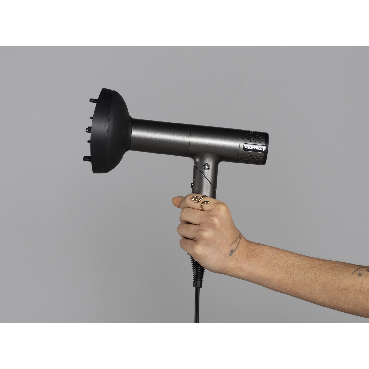 Een hand houdt een slanke, donkergrijze Remington haardroger met diffuser vast tegen een egale grijze achtergrond.