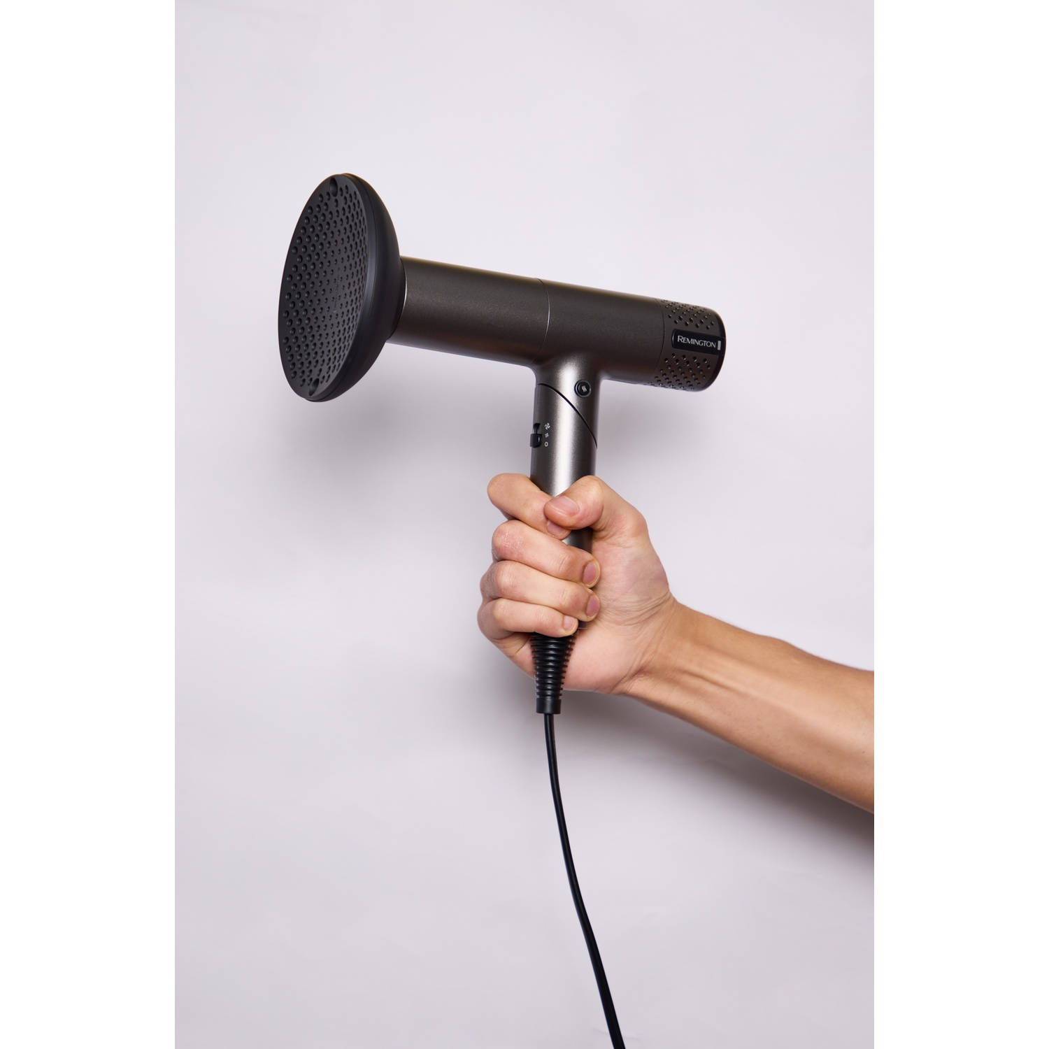Een hand houdt een moderne grijze Remington föhn met diffuser vast tegen een witte achtergrond.